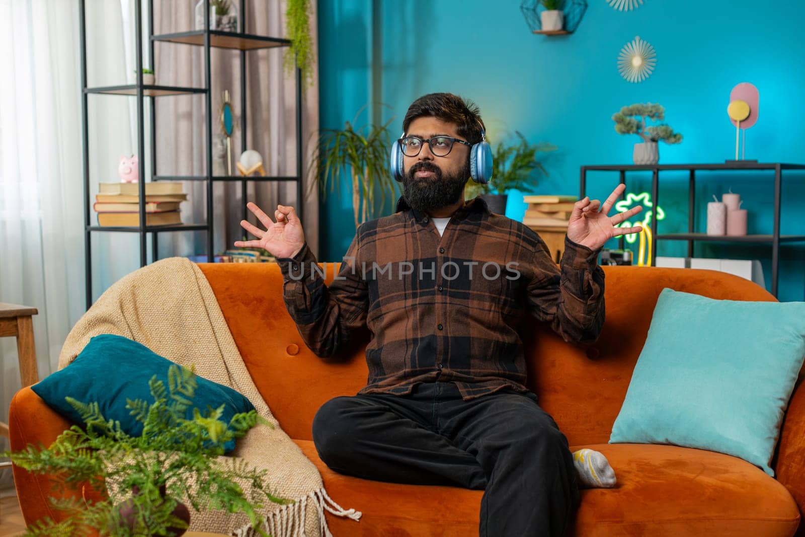 Indian man listening music via headphones, breathes deeply, eyes closed meditating with concentrated thoughts, peaceful mind. Hispanic guy relaxing, taking a break sitting at home living room sofa.