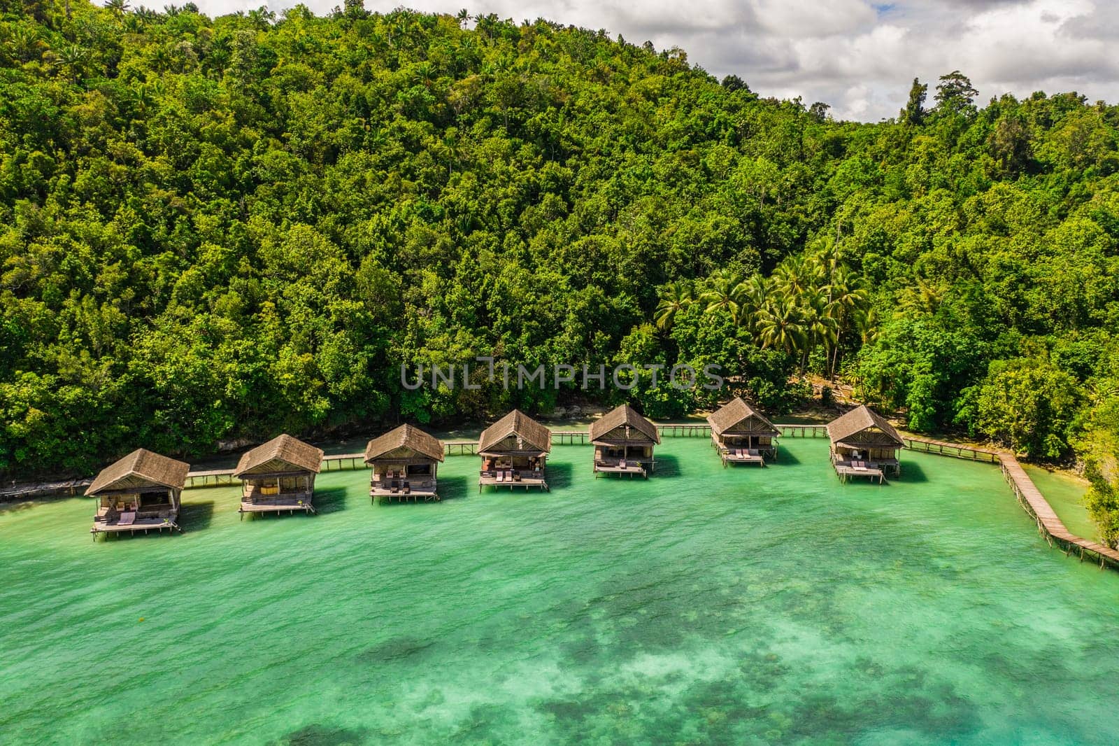 Aerial, bungalow and resort by tropical ocean with hill, island and trees on beach for tourism agency. Jetty, hotel and hospitality by sea, adventure and drone in nature with water in Raja Ampat by YuriArcurs