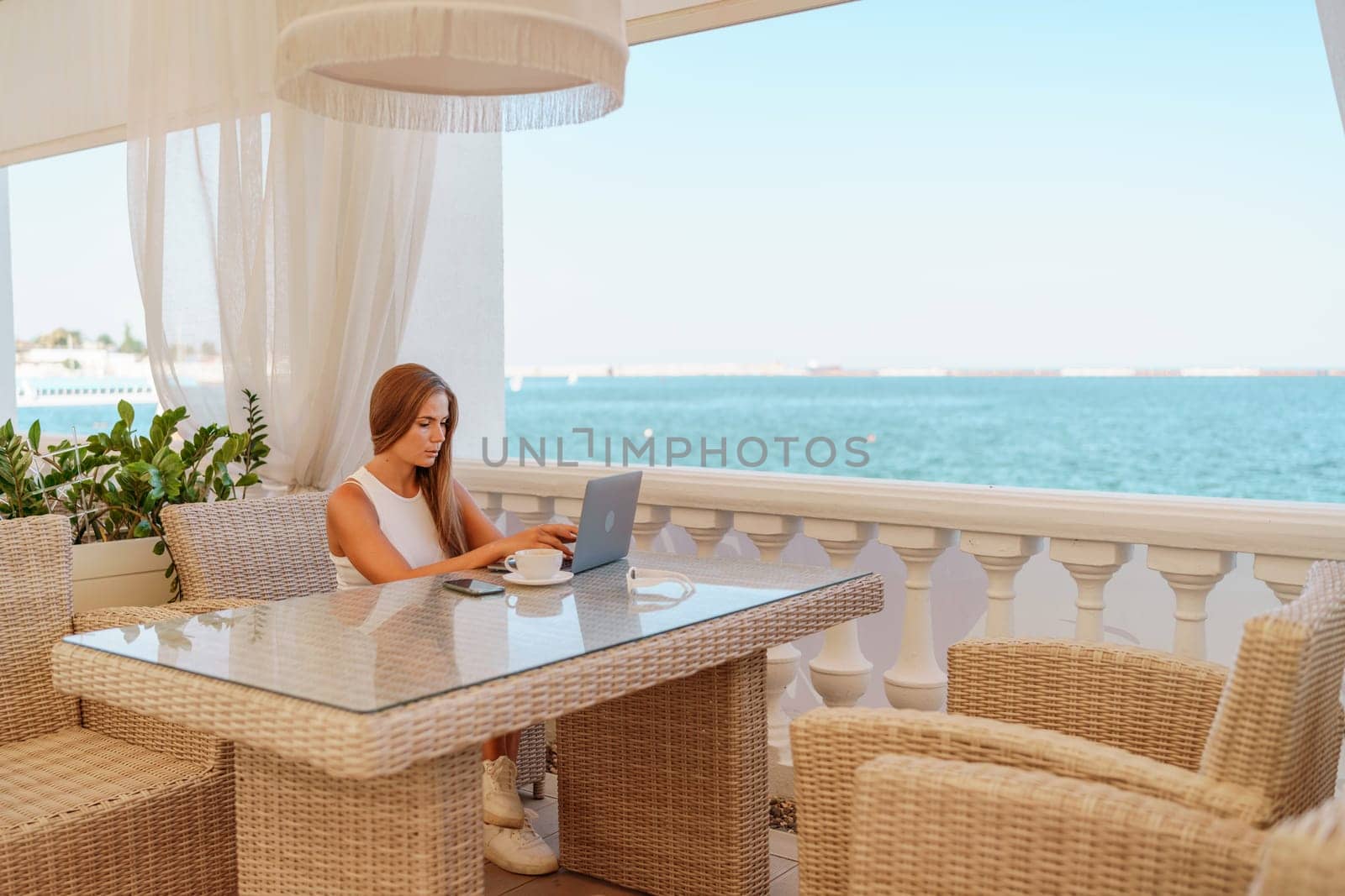 Woman coffee cafe laptop sea. Female freelancer using laptop on coffee break in modern cafe with sea view in the morning.