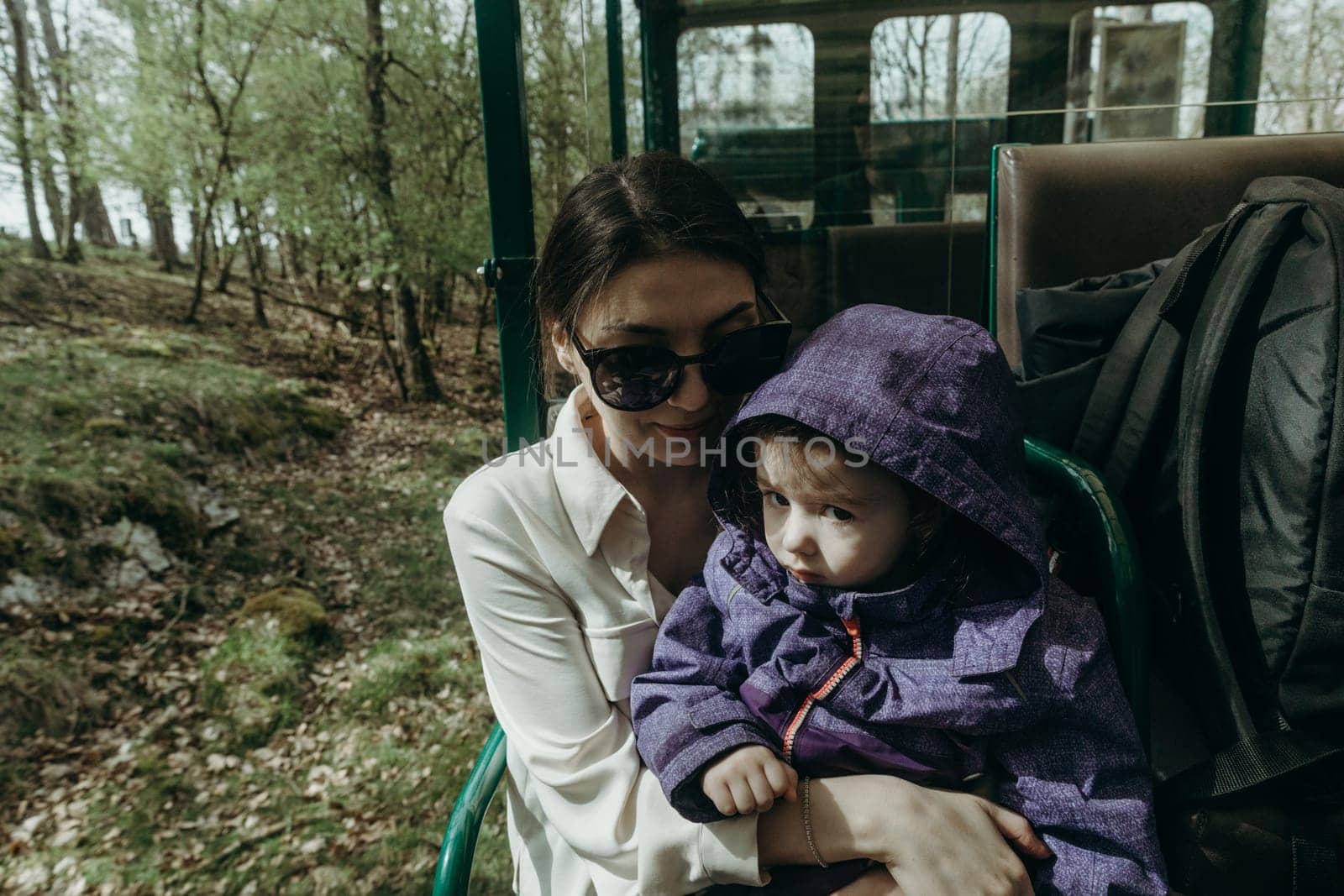 Portrait of one beautiful young Caucasian brunette girl in sunglasses and stylish clothes holds a baby girl in her arms, sitting in an open carriage of a tourist train, driving through a nature reserve on a spring sunny day in a public park, close-up side view.