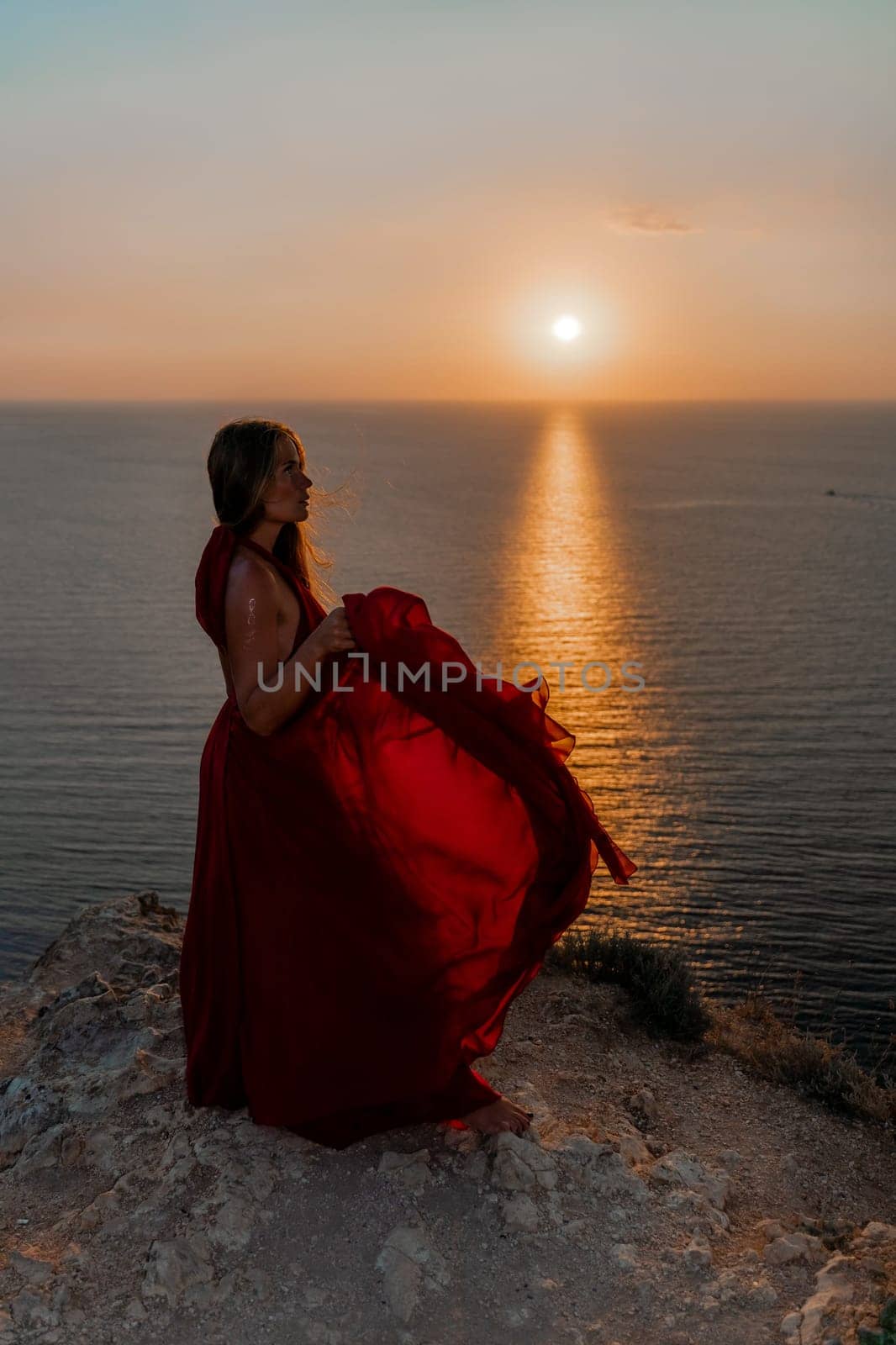 Woman sunset sea red dress, back view a happy beautiful sensual woman in a red long dress posing on a rock high above the sea on sunset