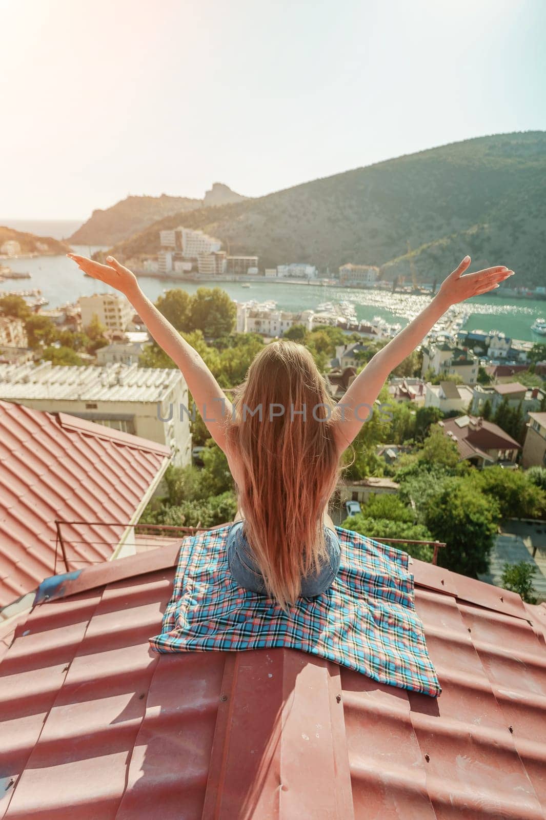 Woman sits on rooftop with outstretched arms, enjoys town view and sea mountains. Peaceful rooftop relaxation. Below her, there is a town with several boats visible in the water by Matiunina