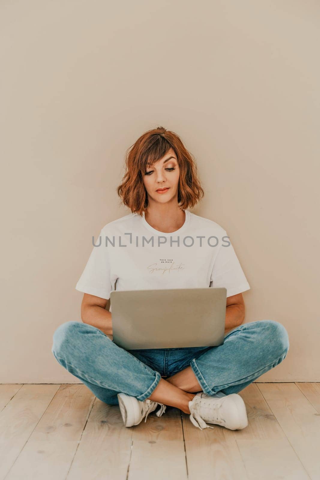 A brunette sits on the floor with a laptop on a beige wall background. She is wearing a white T-shirt, jeans and white sneakers. by Matiunina