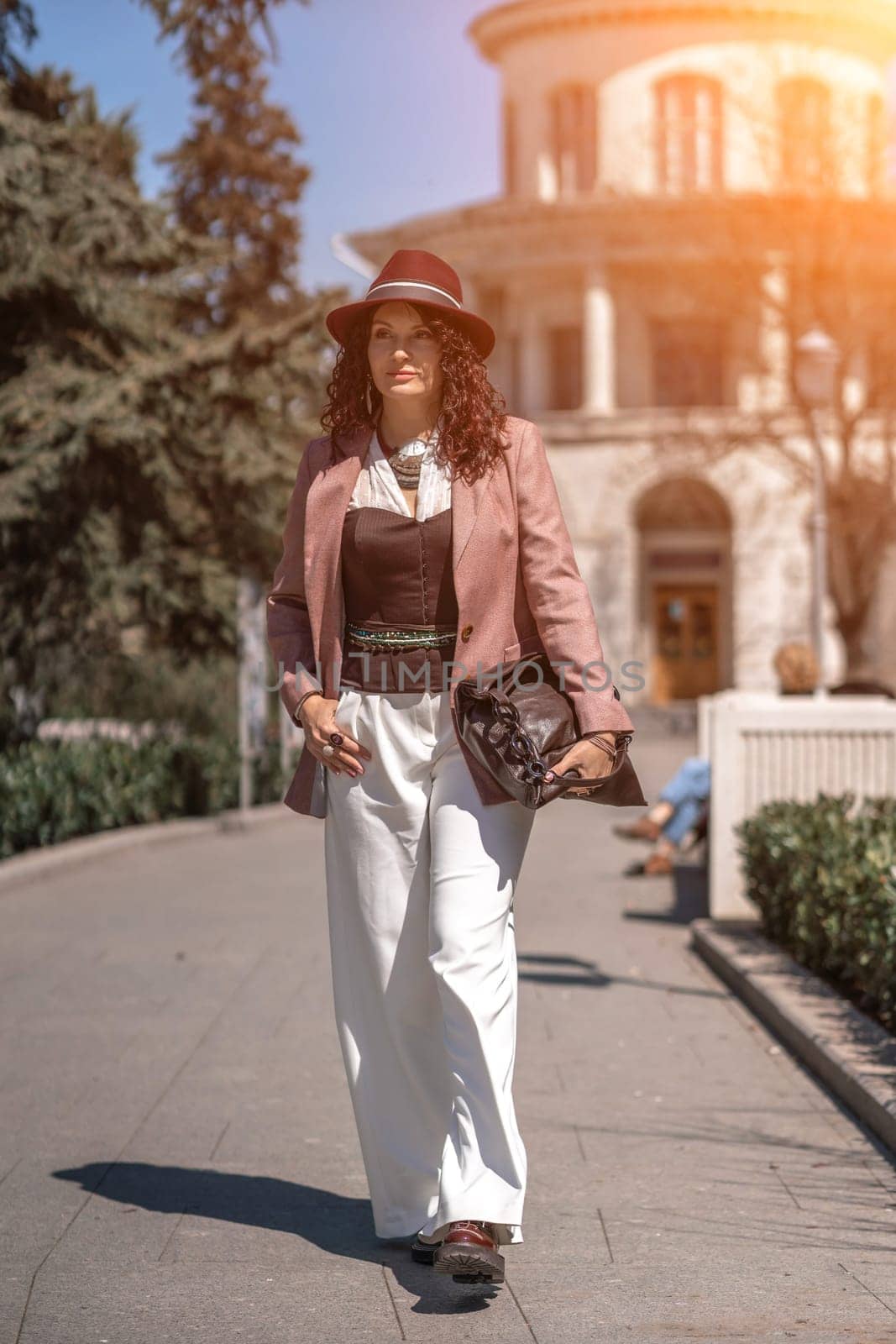 Woman park city. Stylish woman in a hat walks in a park in the city. Dressed in white corset trousers and a pink jacket with a bag in her hands. by Matiunina