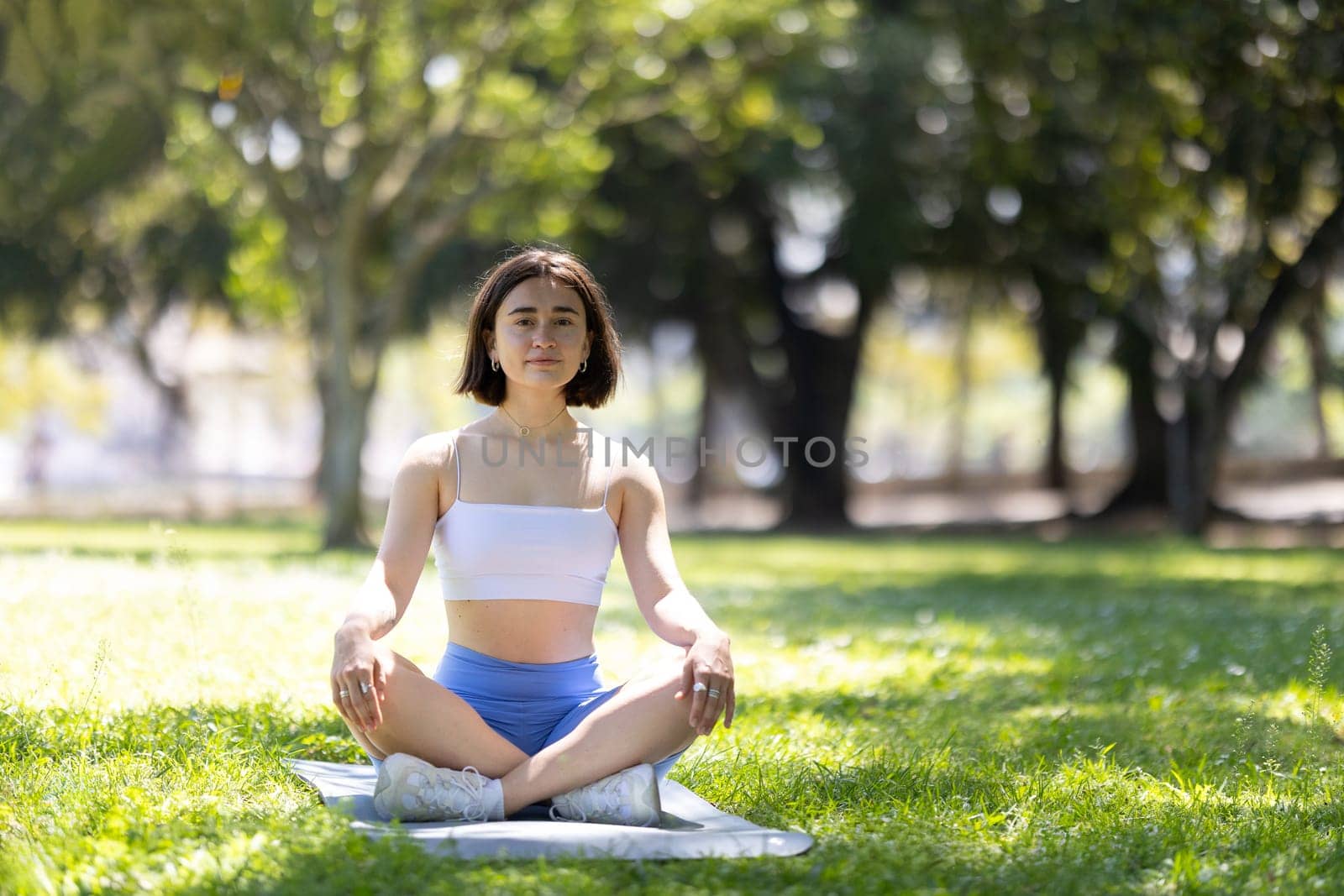 A woman is sitting on a mat in a park, looking up at the sky. She is in a peaceful and relaxed state