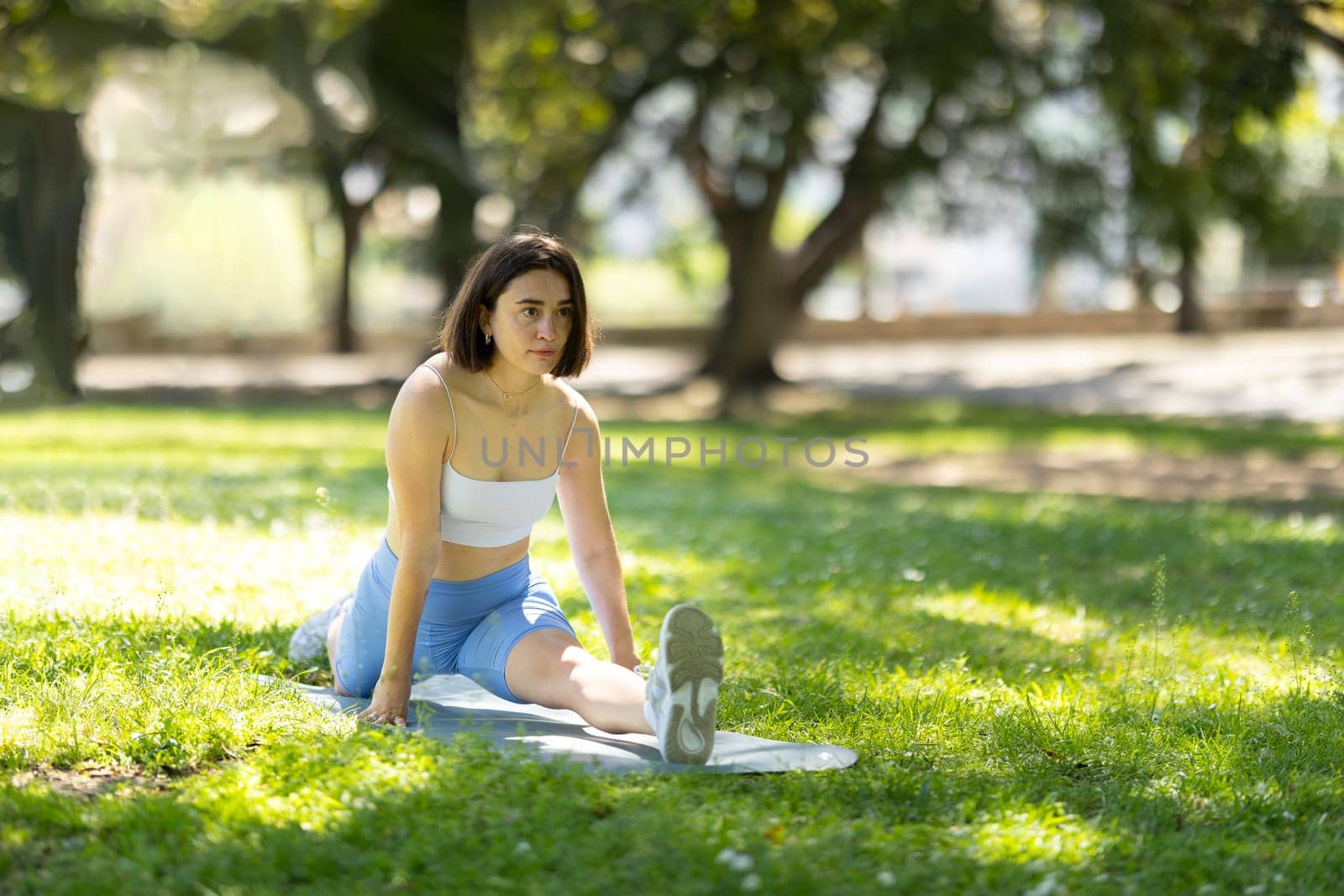 A woman is stretching in a park. She is wearing a white tank top and blue shorts. The grass is green and the sky is blue