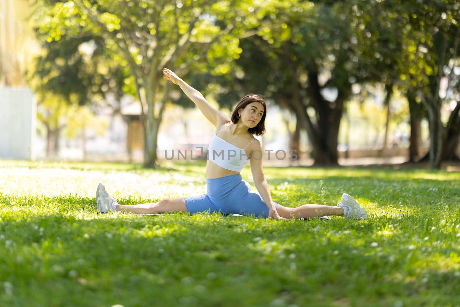 A woman is doing a yoga pose in a park. She is in a split position and is looking up. The scene is peaceful and serene, with the woman's focus on her pose and the surrounding greenery
