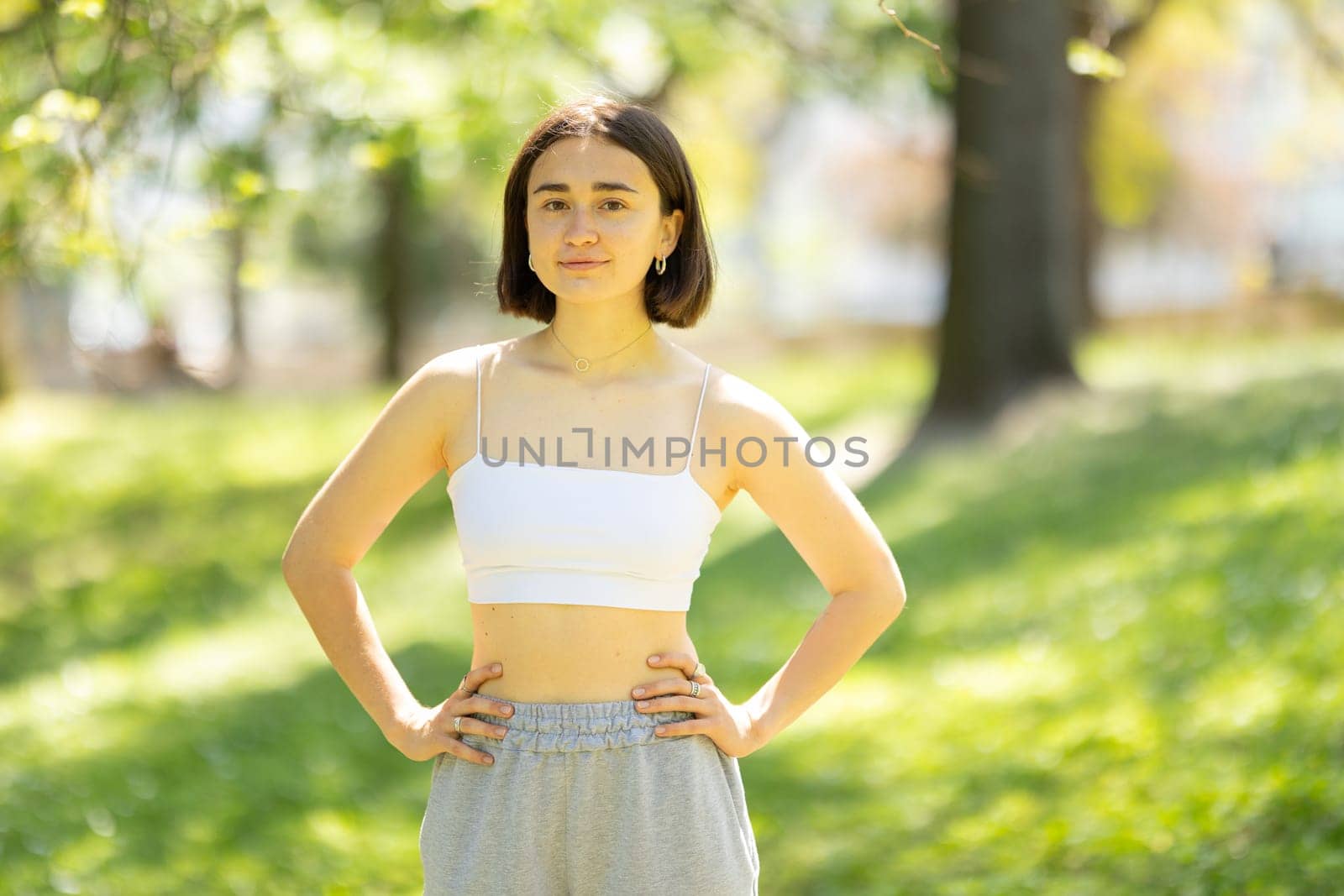 A woman is standing in a park, wearing a white tank top and grey sweatpants. She is posing for a picture, and her body language suggests that she is confident and comfortable in her outfit