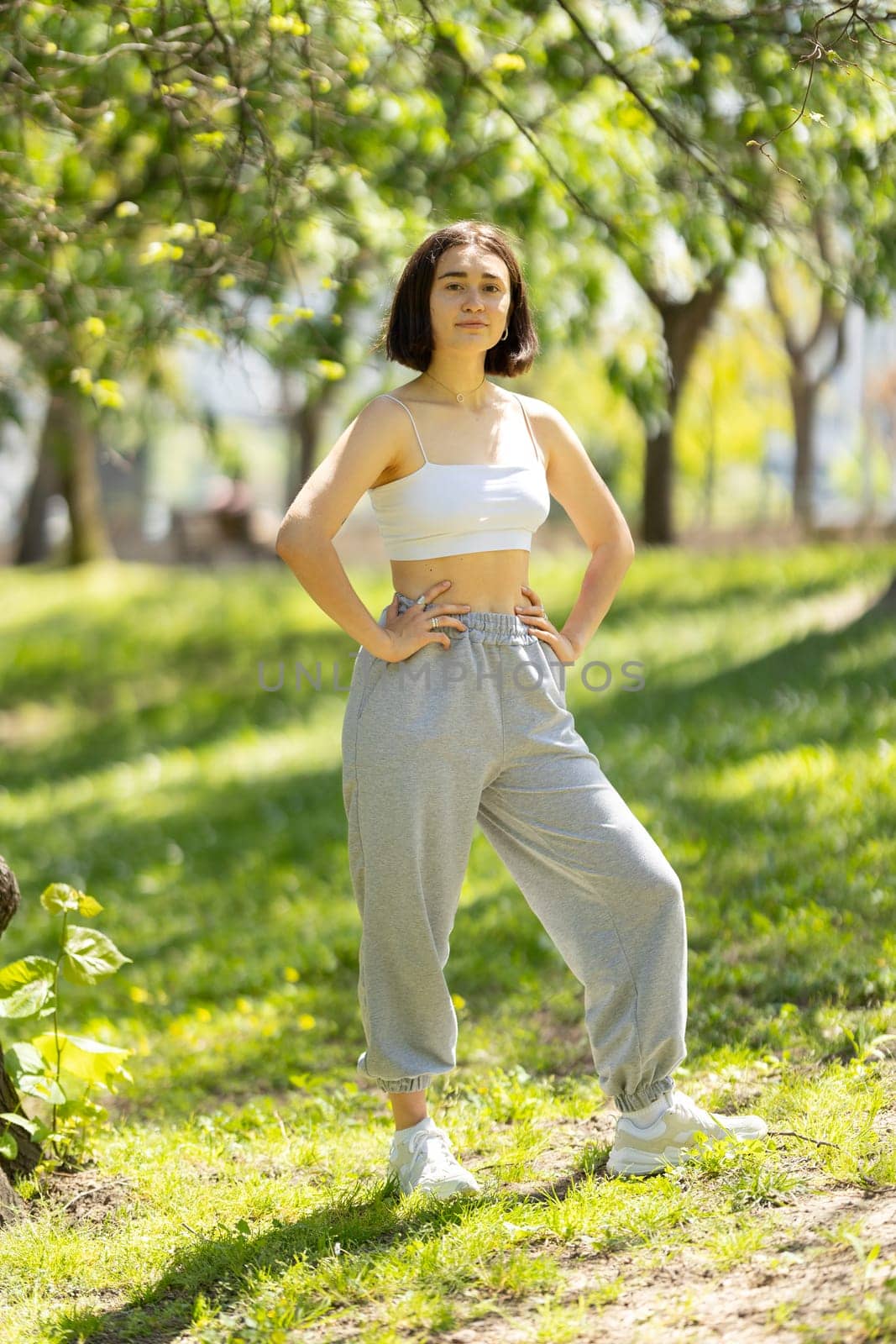 A woman is standing in a park wearing a white tank top and grey sweatpants. She is posing for a picture and she is confident and relaxed