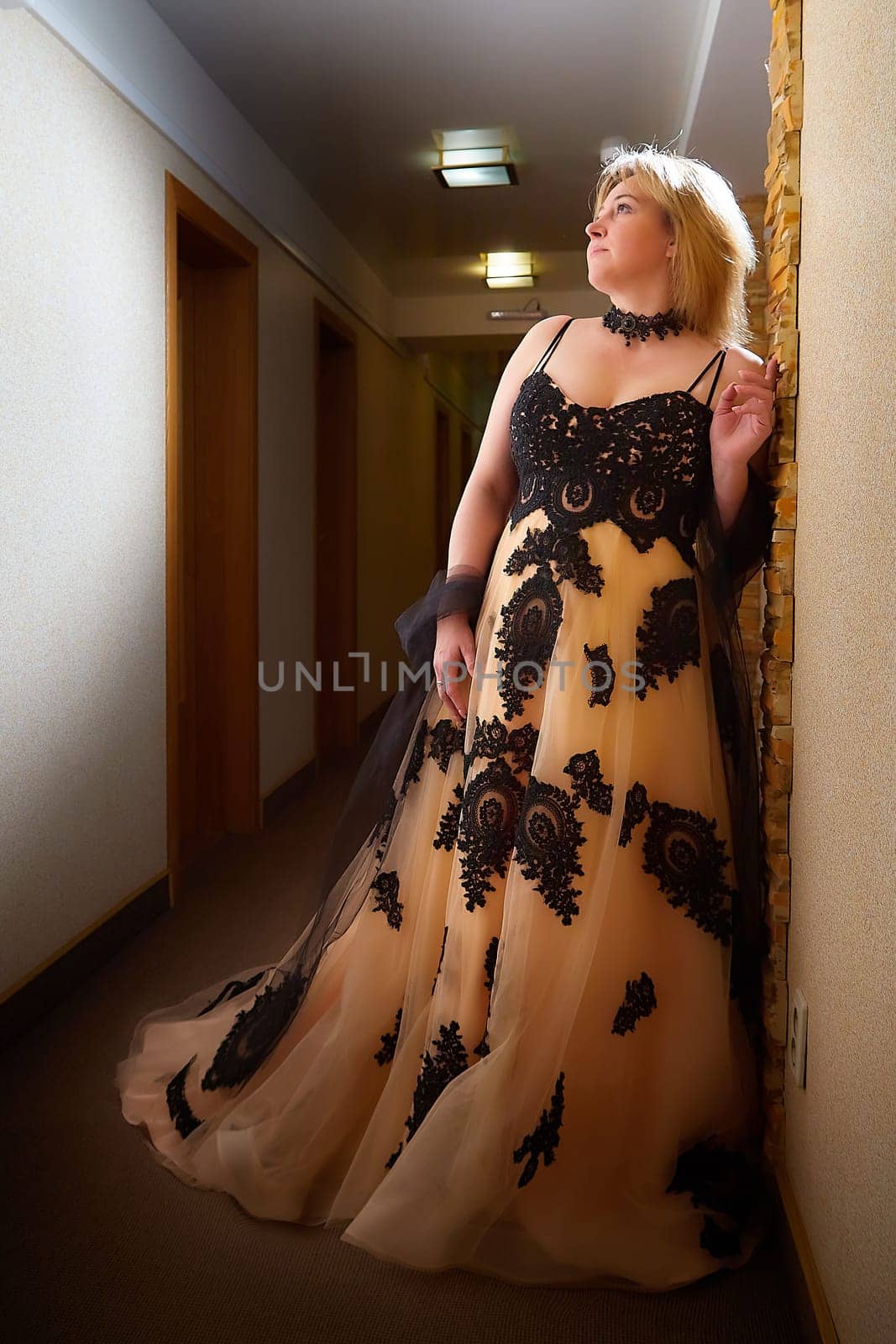 Full length view of a blonde female model, dressed in a black and white queen dress in dark corridor. Mysterious stranger alone inside castle or hotel