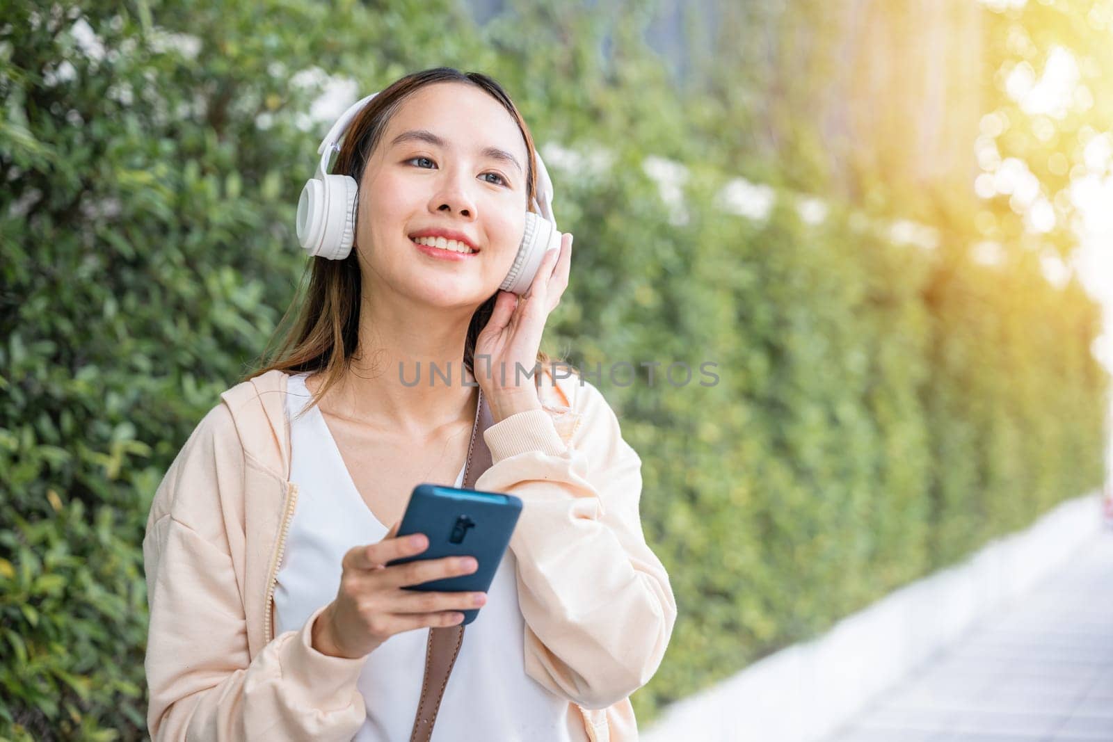 In midst of an urban city garden a happy young woman uses wireless headphones to choose and enjoy her favorite music dancing with pure joy. Her carefree spirit is a beautiful reflection of season. by Sorapop