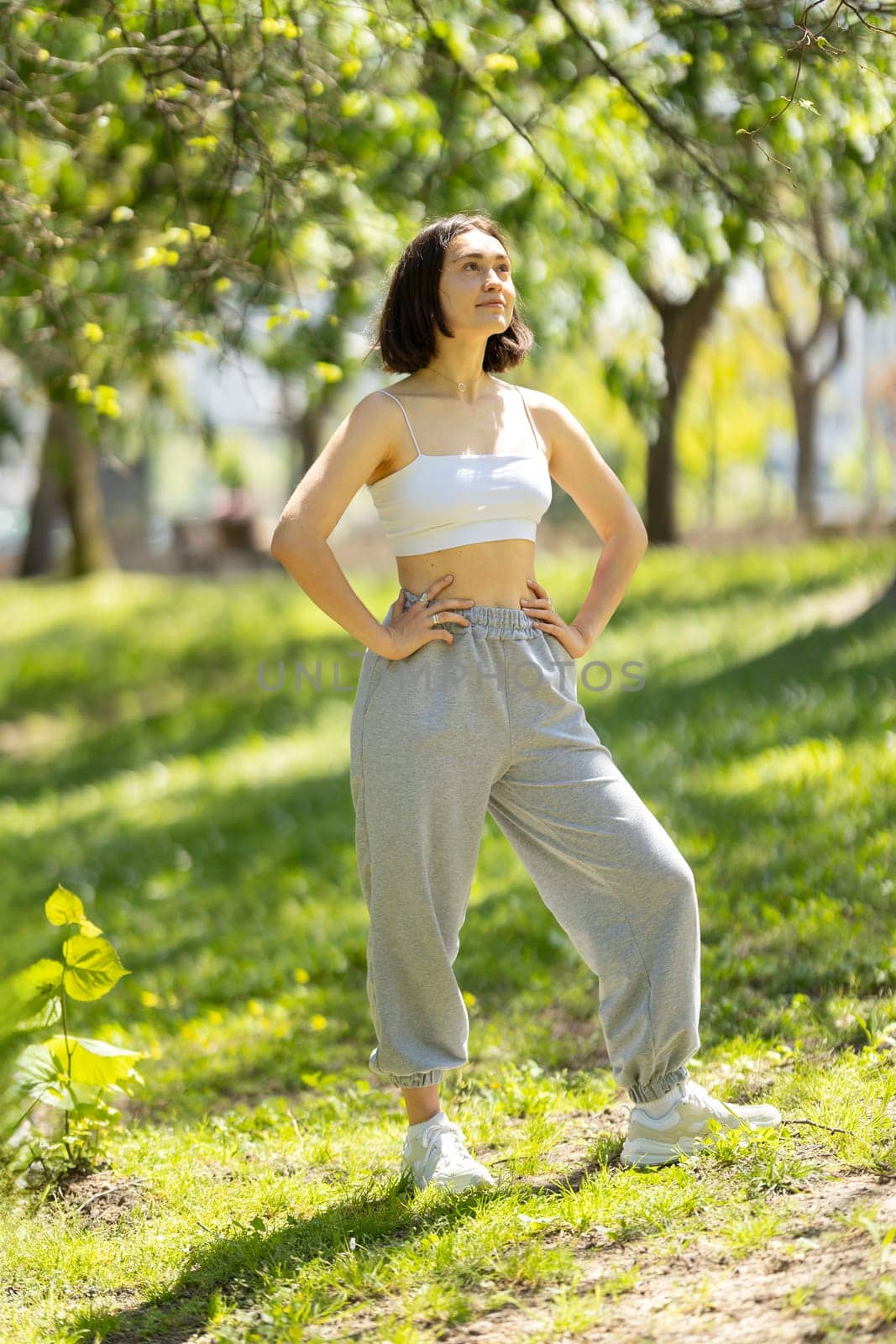A woman is standing in a park, wearing a white tank top and grey sweatpants by Studia72