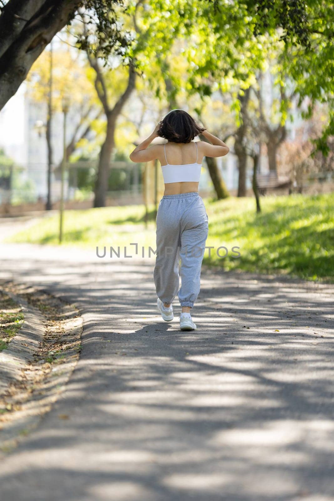 A woman is walking down a path in a park. She is wearing headphones and has her hands on her head. The scene is peaceful and serene, with the woman enjoying her walk in the park