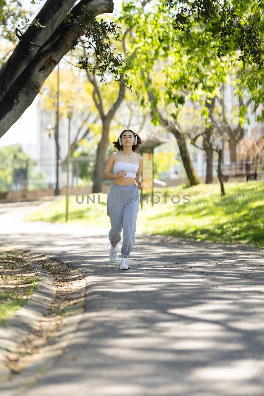 A woman is running on a path in a park. She is wearing headphones and a white tank top