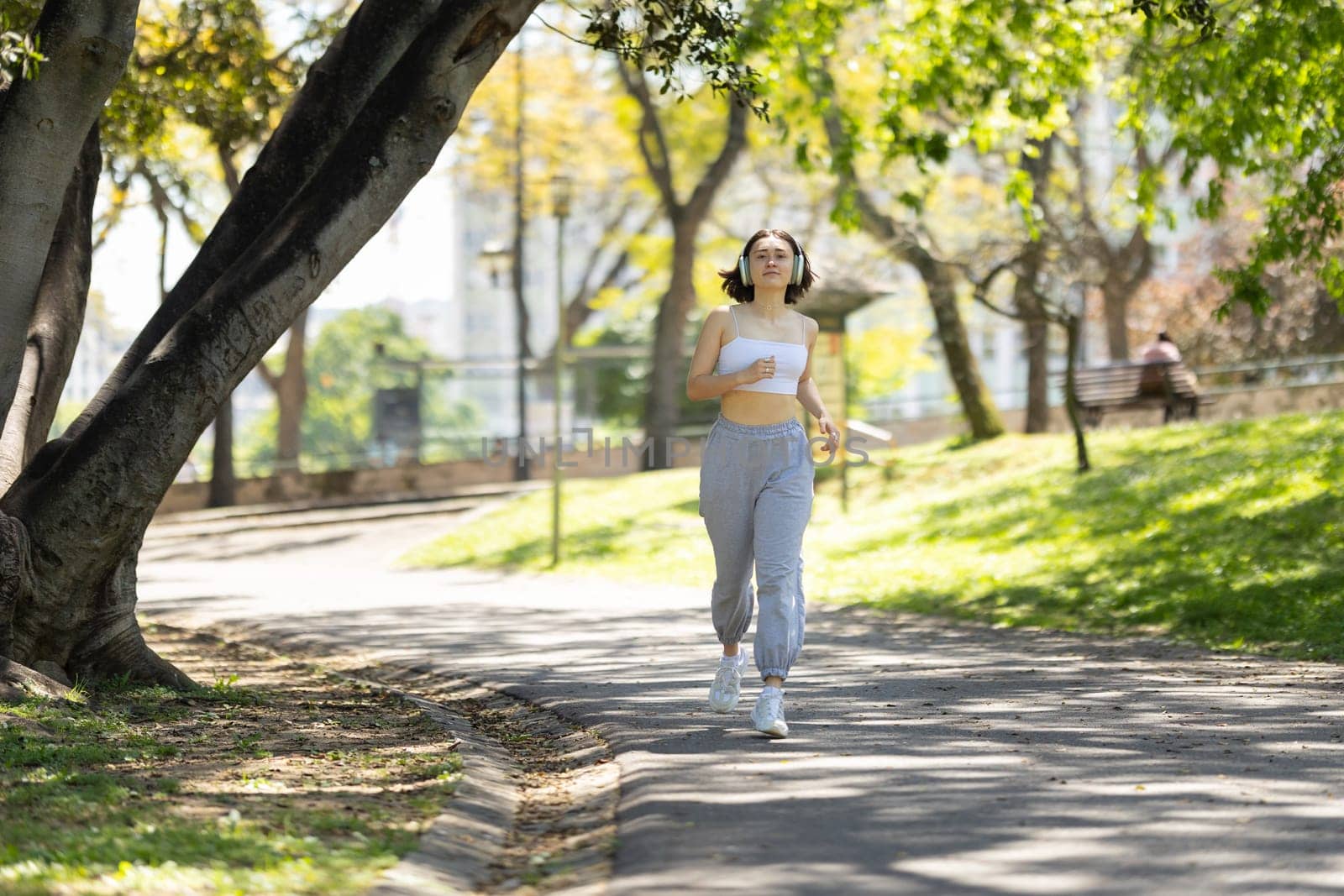 A woman runs on a path in a park. She is wearing headphones and a white tank top