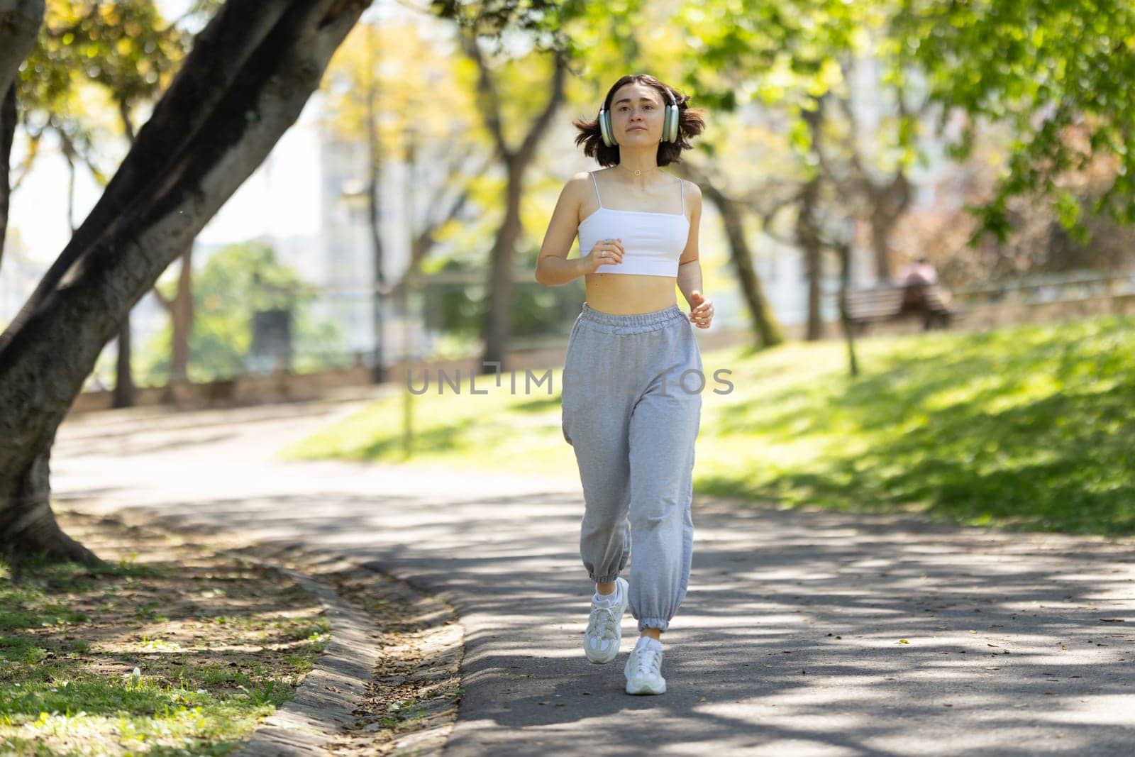 A woman is running in a park with headphones on. She is wearing a white tank top and gray pants