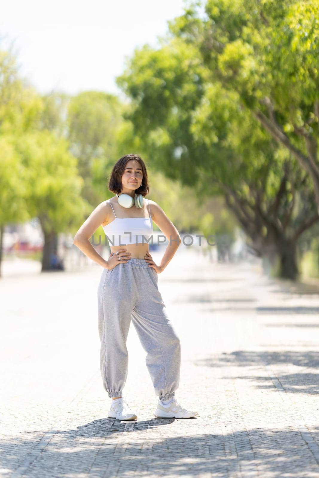 A woman is standing on a sidewalk wearing headphones and a white tank top by Studia72