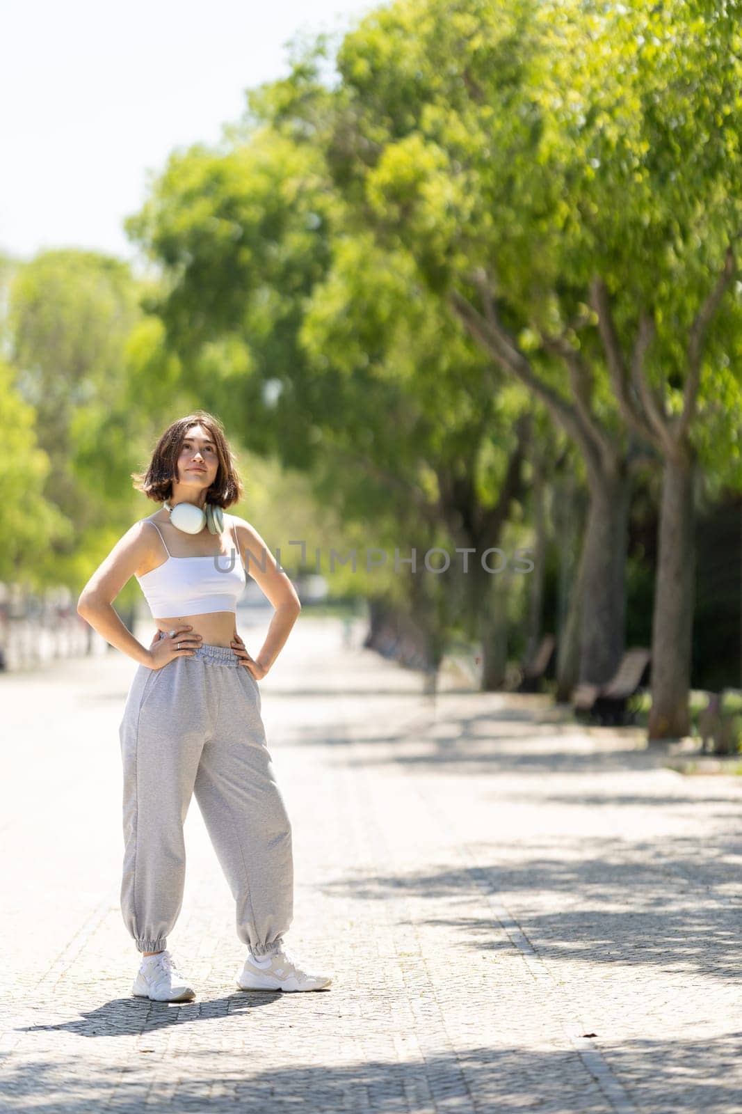 A woman is standing on a sidewalk in front of trees. She is wearing a white tank top and gray pants