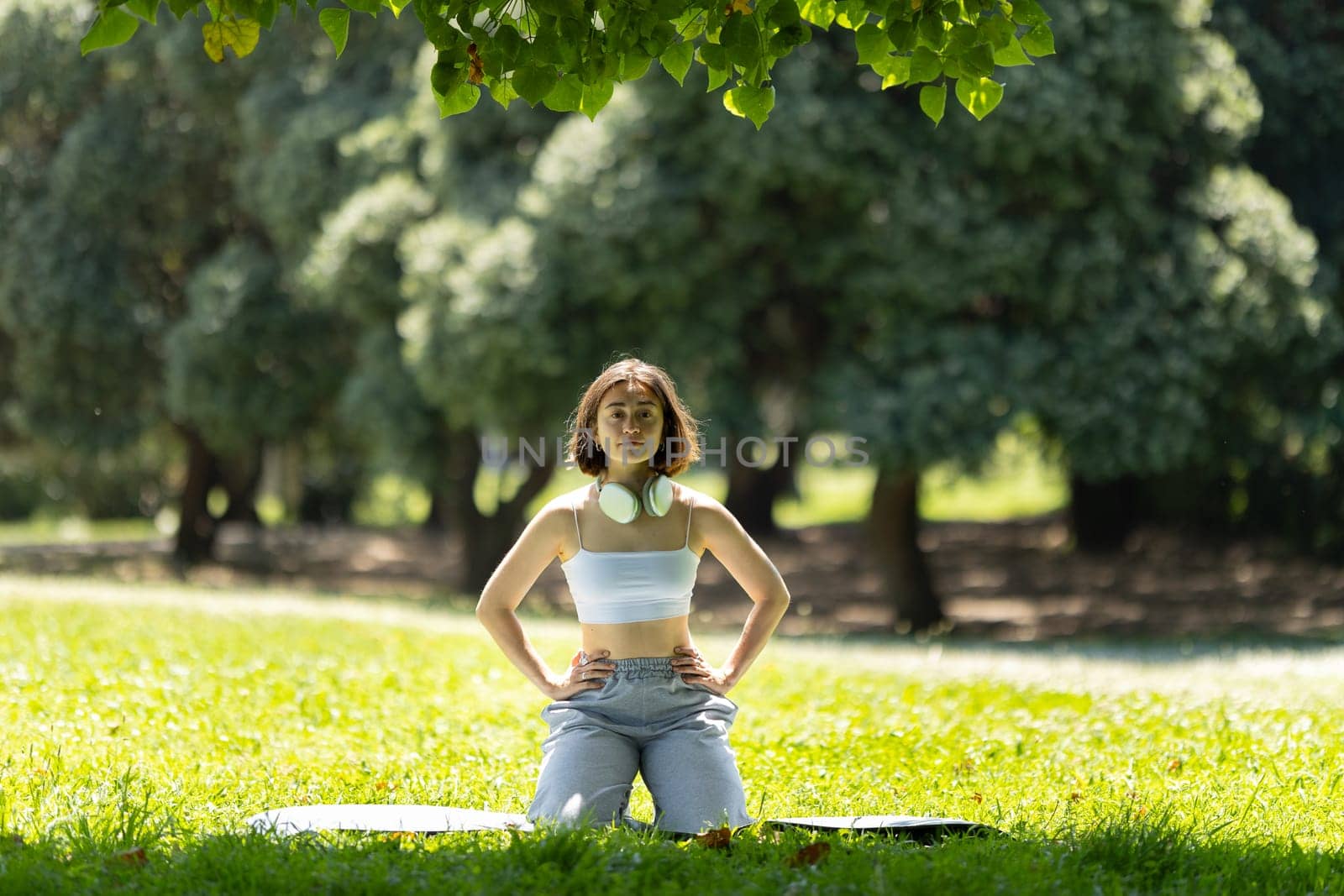 A woman is sitting on the grass in a park. She is wearing headphones and has her arms crossed