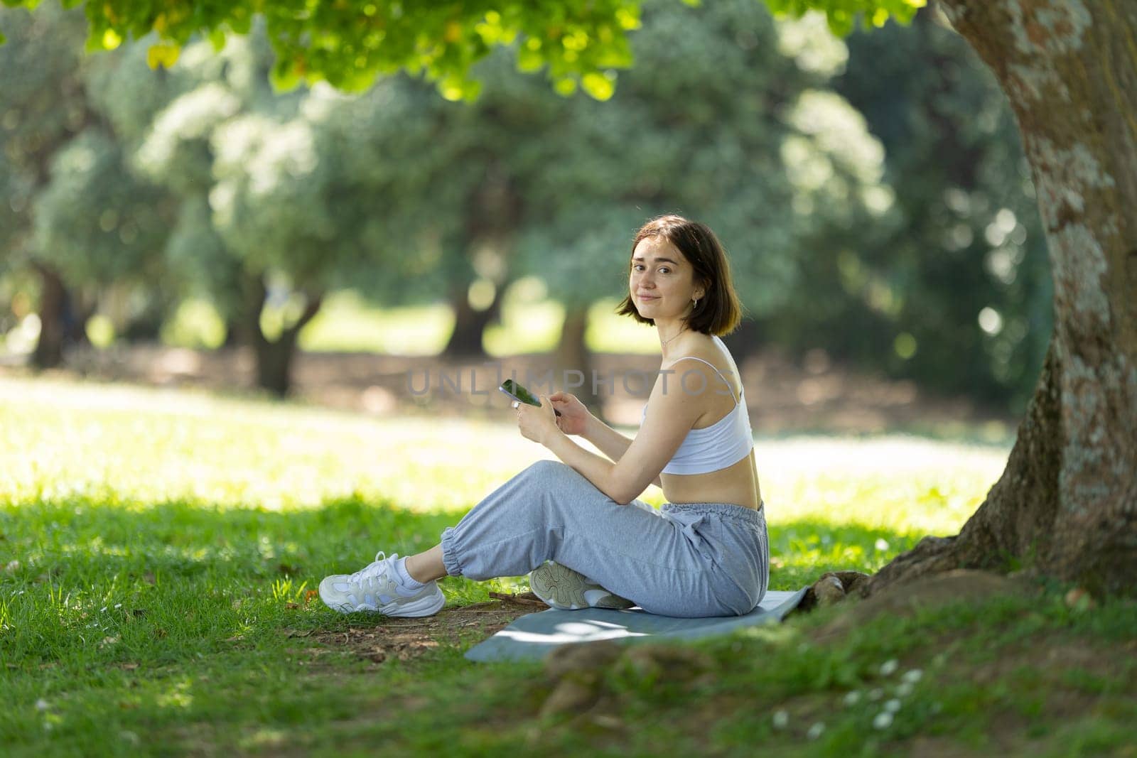 A sportive woman is sitting under a tree and use phone. She is wearing a white tank top and gray pants
