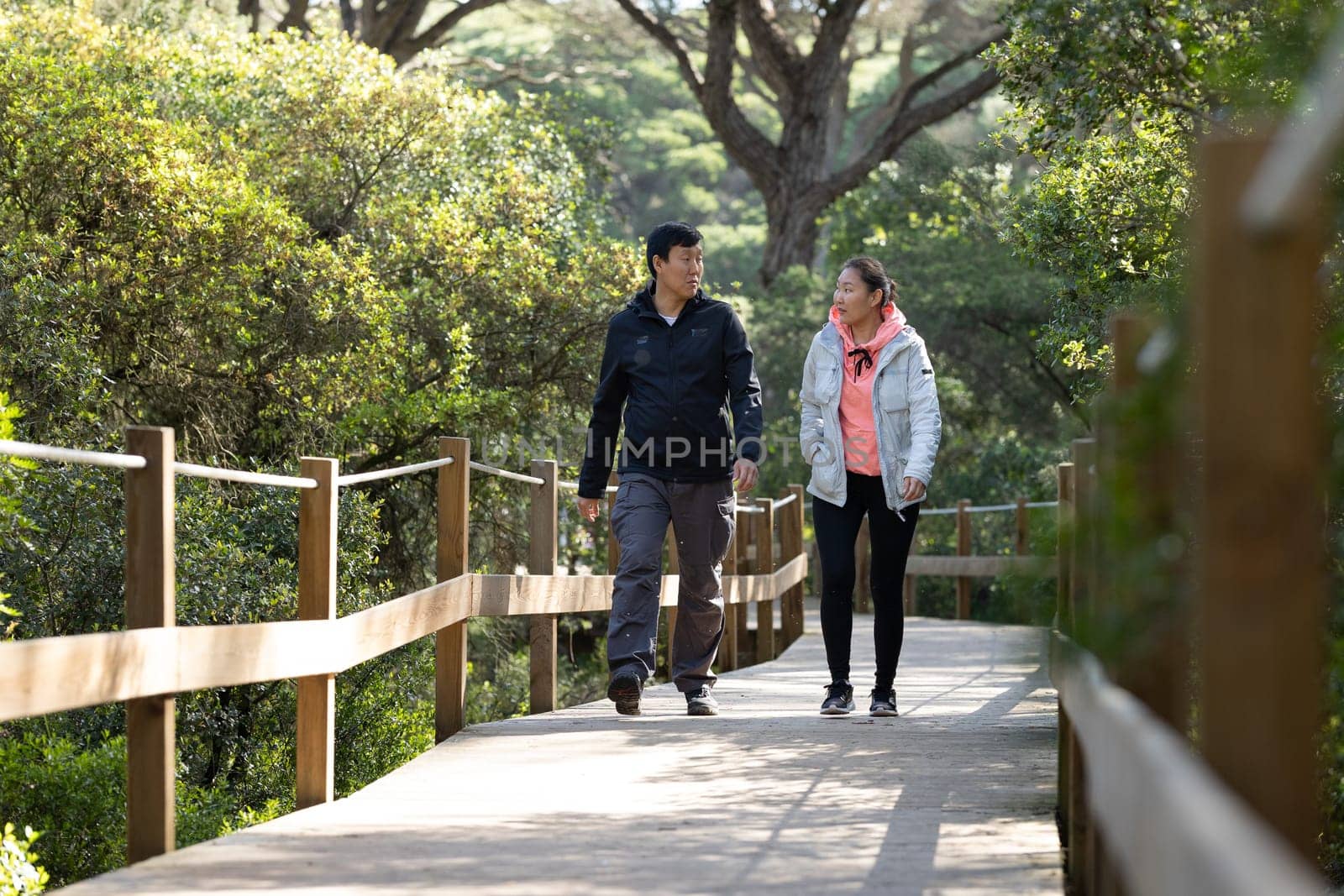 A man and woman are walking on a wooden bridge. The man is wearing a black jacket and the woman is wearing a pink jacket. They are holding hands and seem to be enjoying each other's company