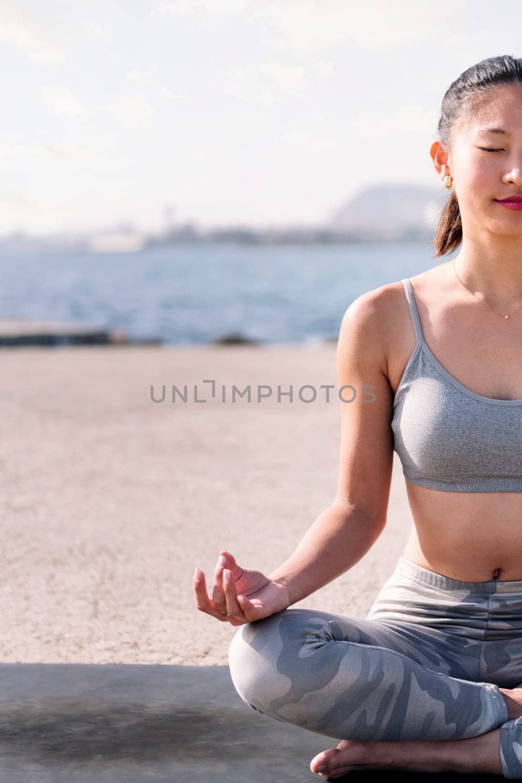 young asian woman in sportswear doing meditation by the sea sitting with legs crossed, concept of mental relaxation and healthy lifestyle, copy space for text