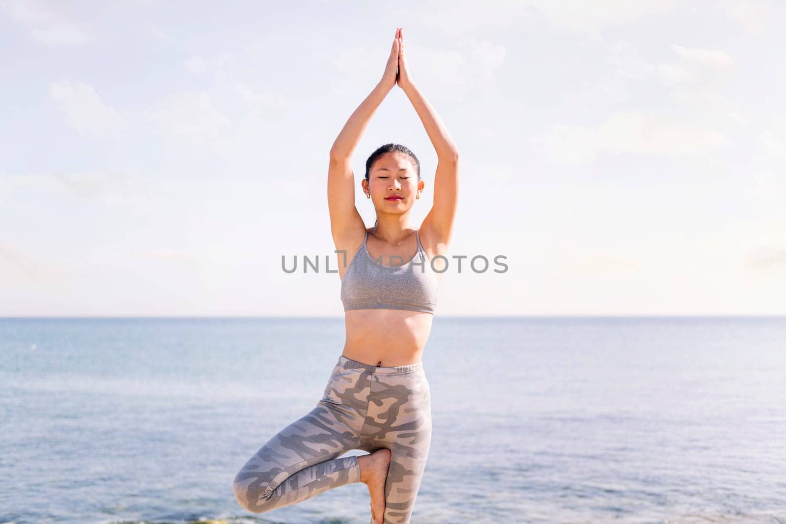 young asian woman in sportswear doing yoga tree position at beach, concept of mental relaxation and healthy lifestyle