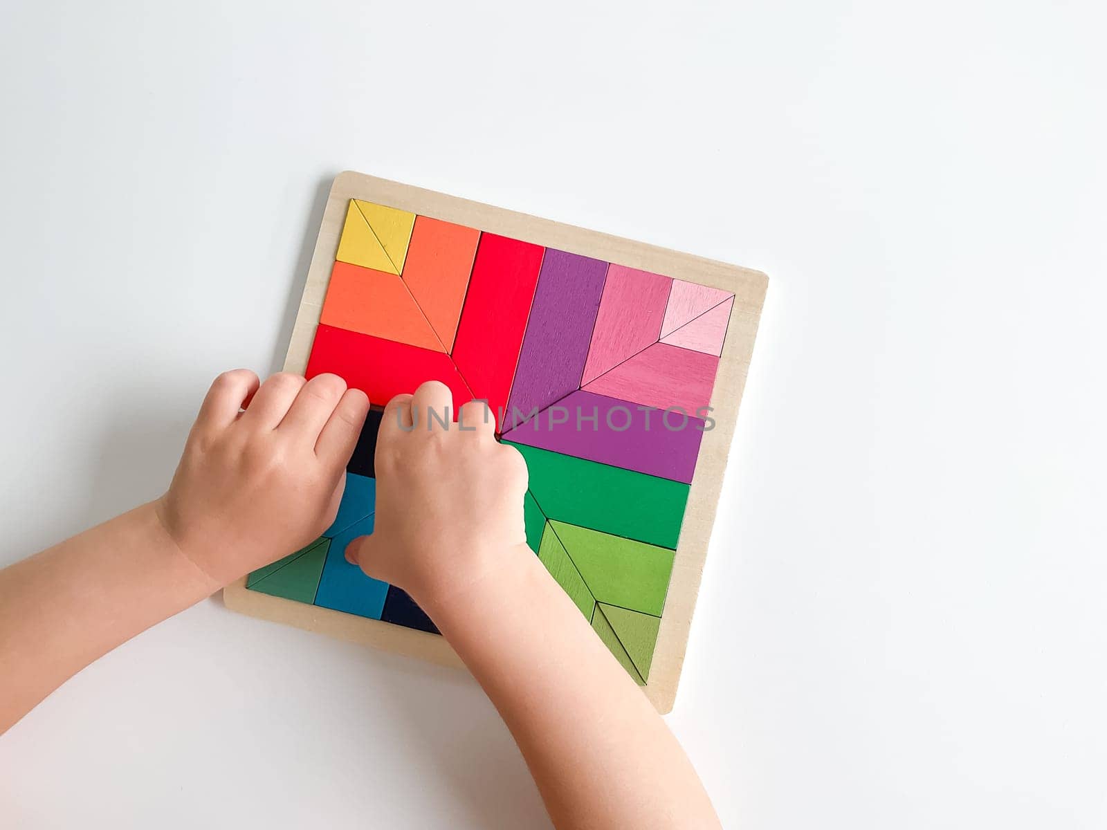 child's hand collects multicolored wooden mosaic on white background. child solves a colorful tangram. square of colorful geometric shapes on white background.