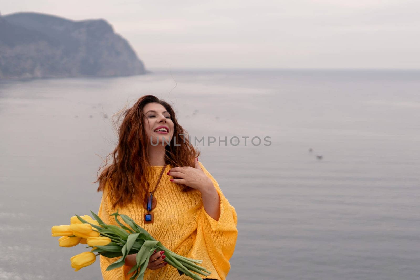 Portrait happy woman woman with long hair against a background of mountains and sea. Holding a bouquet of yellow tulips in her hands, wearing a yellow sweater.