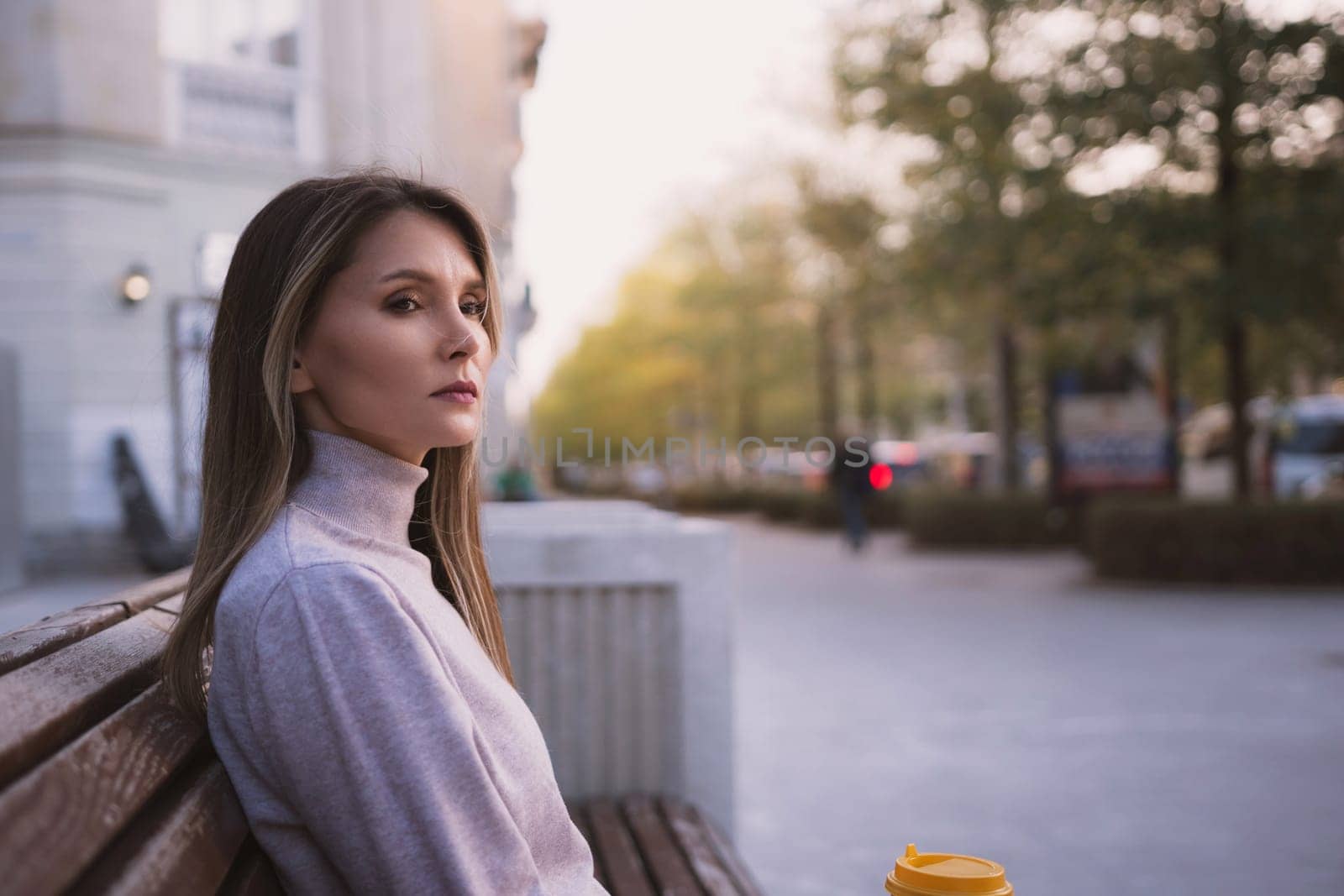 A woman sits on a bench in a city street, looking at the camera. She is wearing a gray sweater and has a cup in her hand. The scene is quiet and peaceful