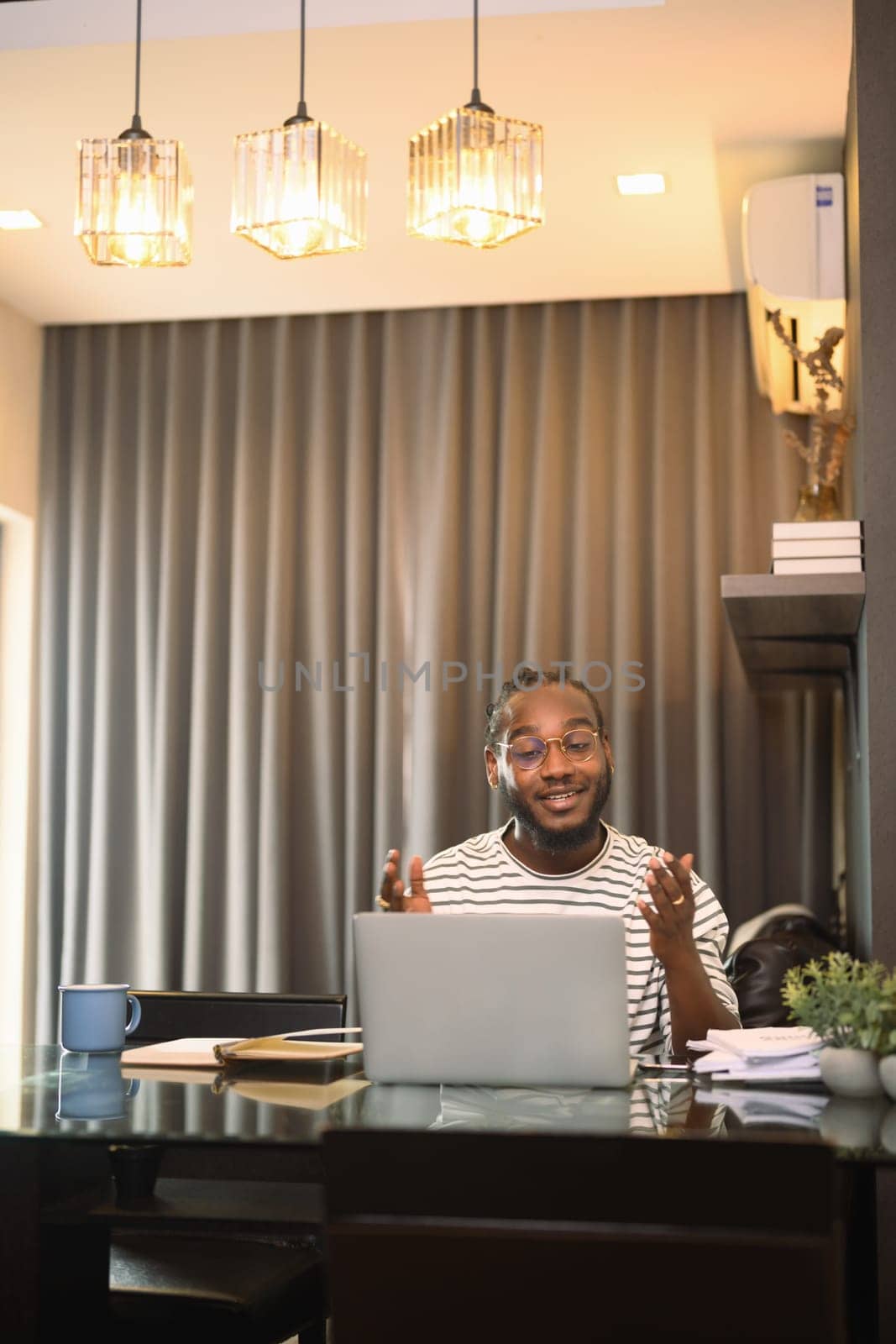 Cheerful African American man having video call on laptop sitting at table in kitchen by prathanchorruangsak