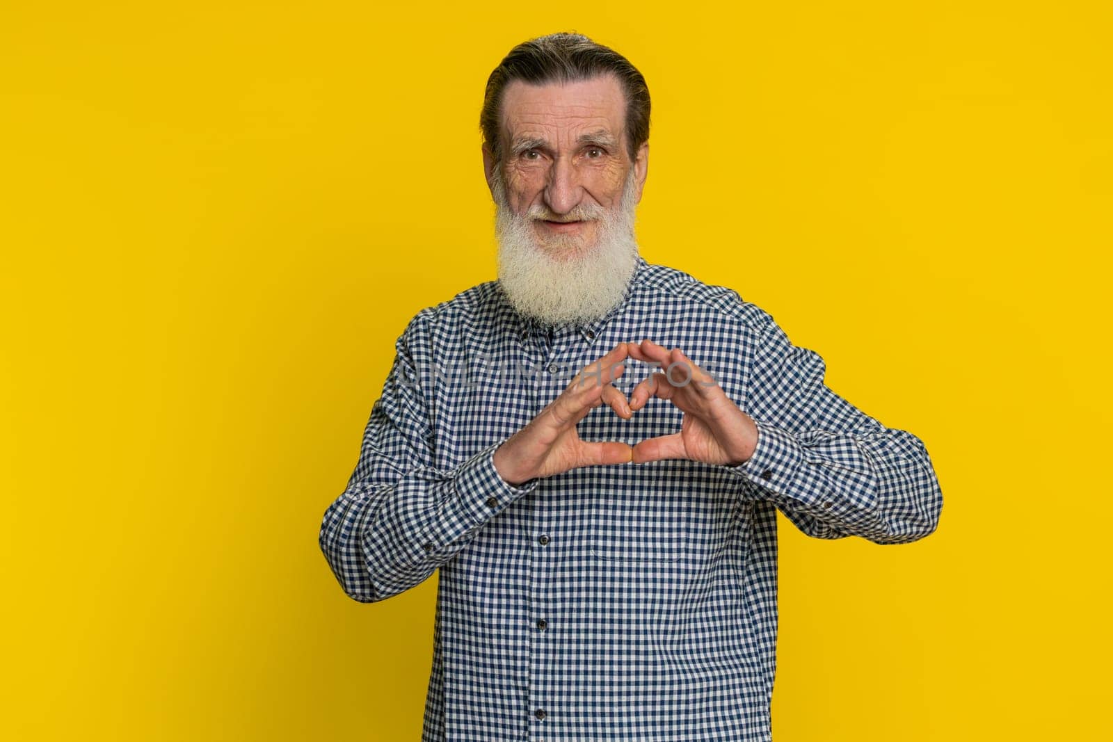 Man in love. Smiling attractive senior old man makes heart gesture demonstrates love sign expresses good positive feelings and sympathy. Elderly bearded grandfather isolated on yellow background