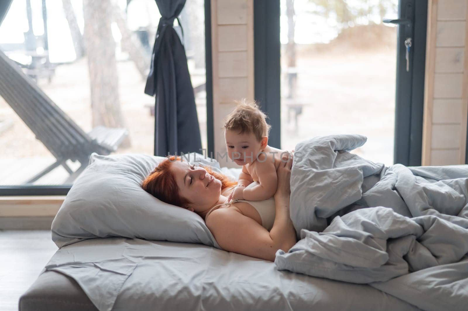 Red-haired Caucasian woman lies in bed with her baby son in a country house with panoramic windows. by mrwed54