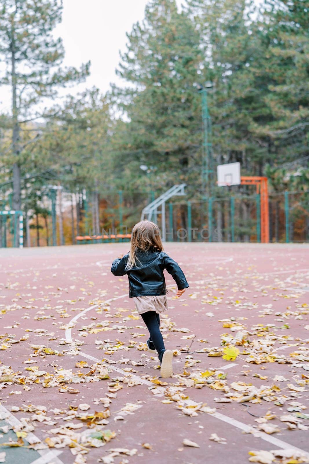 Little girl runs along a basketball court strewn with fallen leaves in the park. Back view. High quality photo