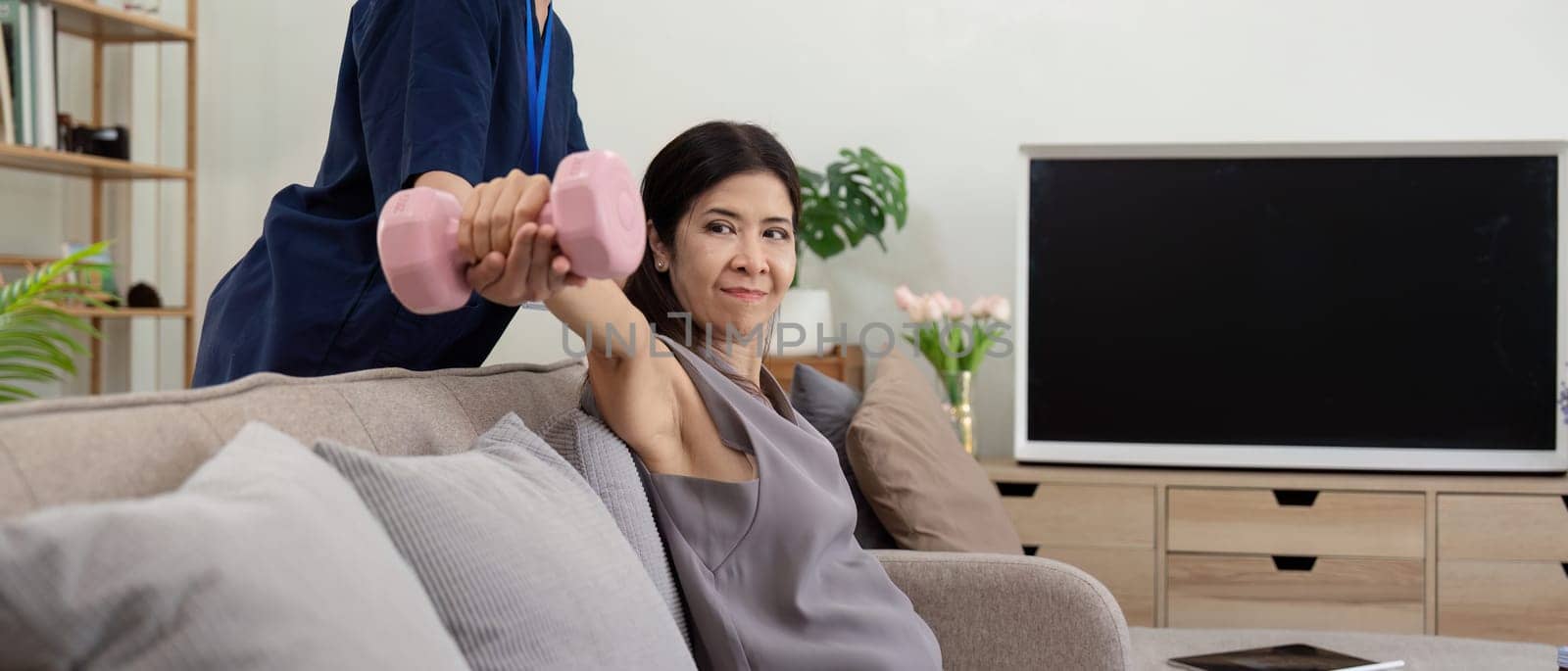 Old woman training with physiotherapist using dumbbells at home. Therapist asian assisting senior woman with exercise in nurse home. Elderly patient using dumbbells with outstretched arms.