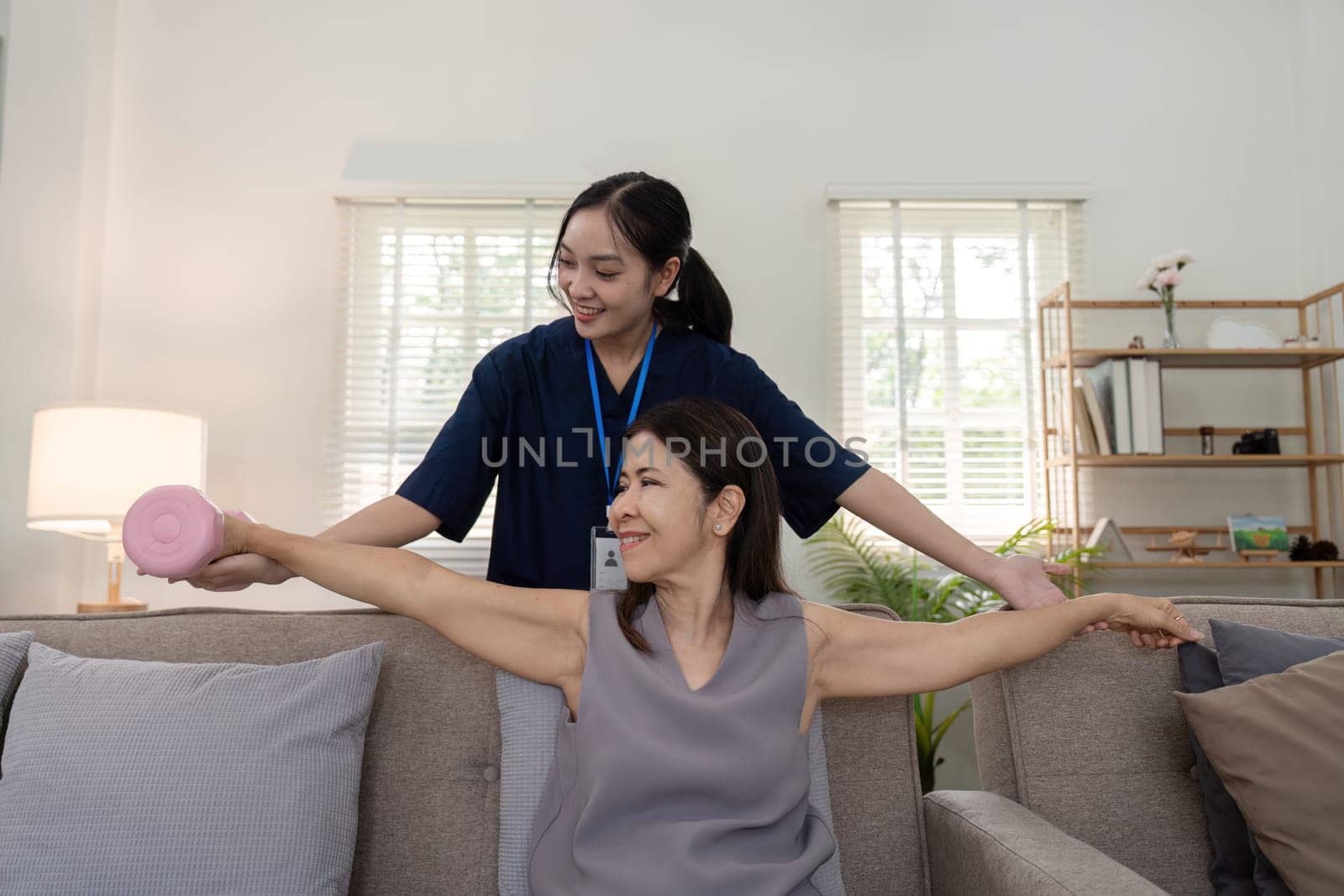Old woman training with physiotherapist using dumbbells at home. Therapist asian assisting senior woman with exercise in nurse home. Elderly patient using dumbbells with outstretched arms.