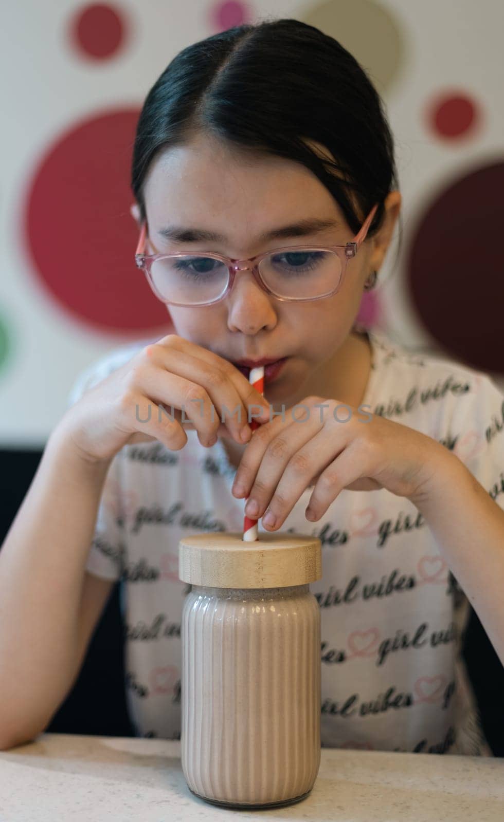 Portrait of one beautiful caucasian girl drinking through a straw of chocolate millshake from a glass sitting at a table on a sofa in a cafe against the background of an abstract wall, close-up side view.