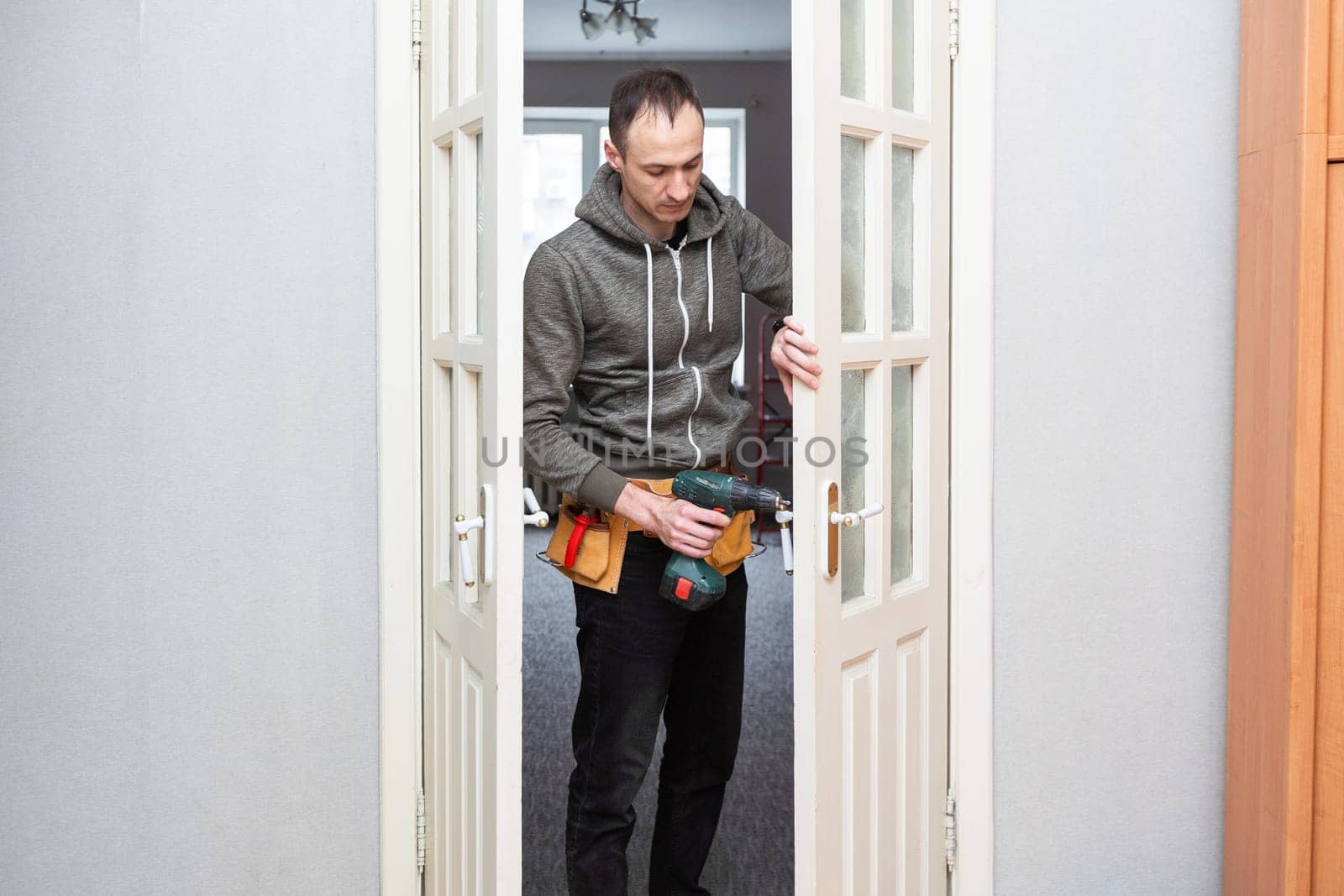 Maintenance Concept. Focused serious young locksmith with tattoo on hand wearing blue uniform standing on one knee in doorway installing doorknob handle, repairing wooden front door entrance. High quality photo
