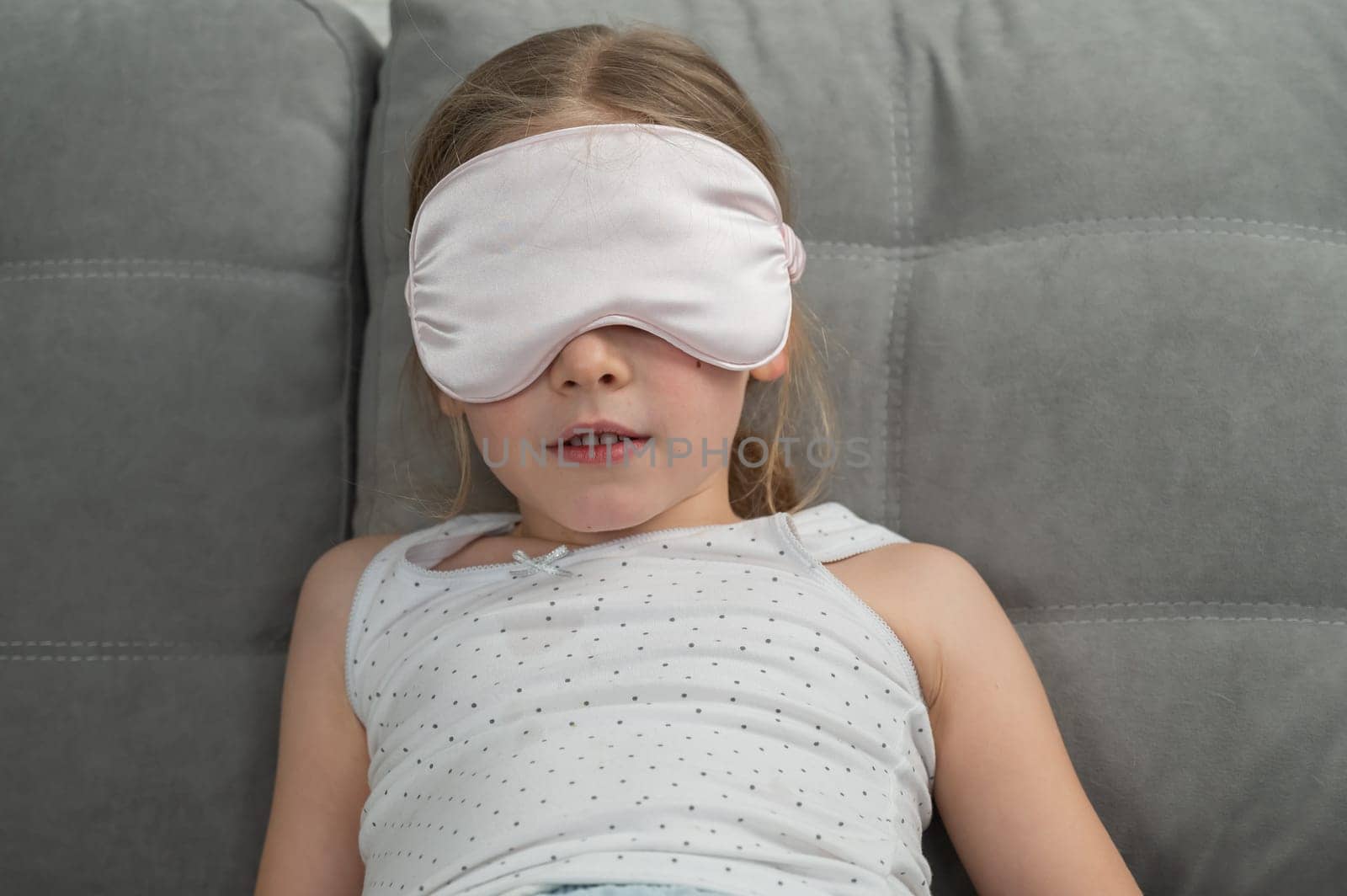 Little Caucasian girl puts on a sleep mask while sitting on the sofa. Doesn't want to sleep. by mrwed54