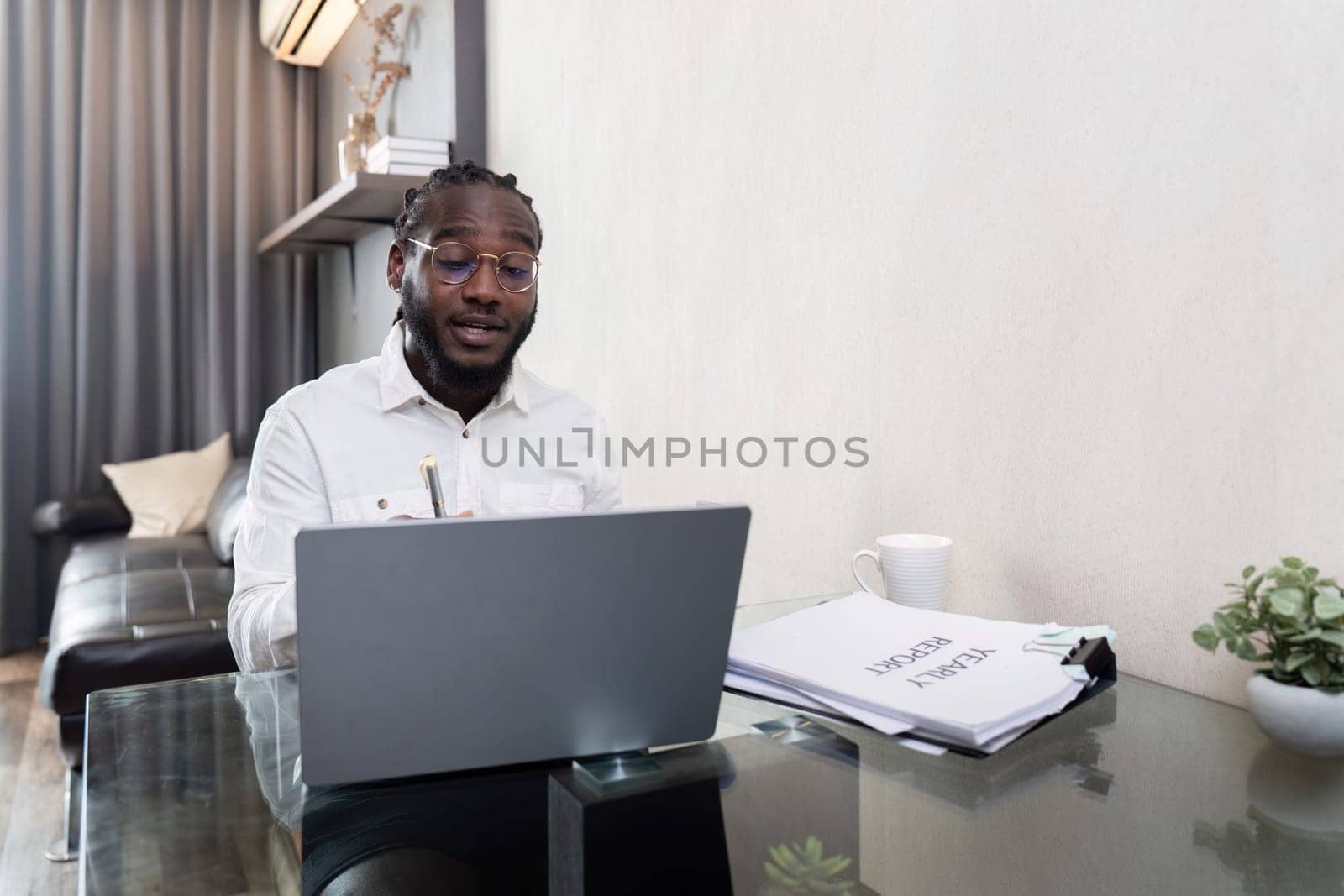 African American man working with laptop computer remote while sitting at glass table in living room. Black guy do freelance work at home office by nateemee