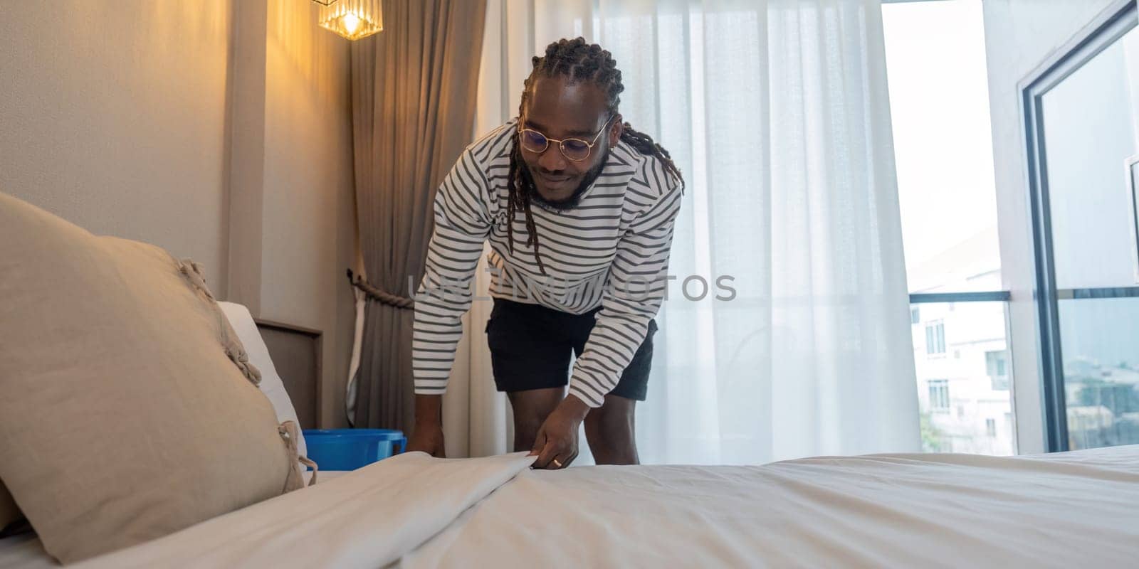 Young African American man clean making bed. happy black man changing bedsheets at home standing in bedroom.