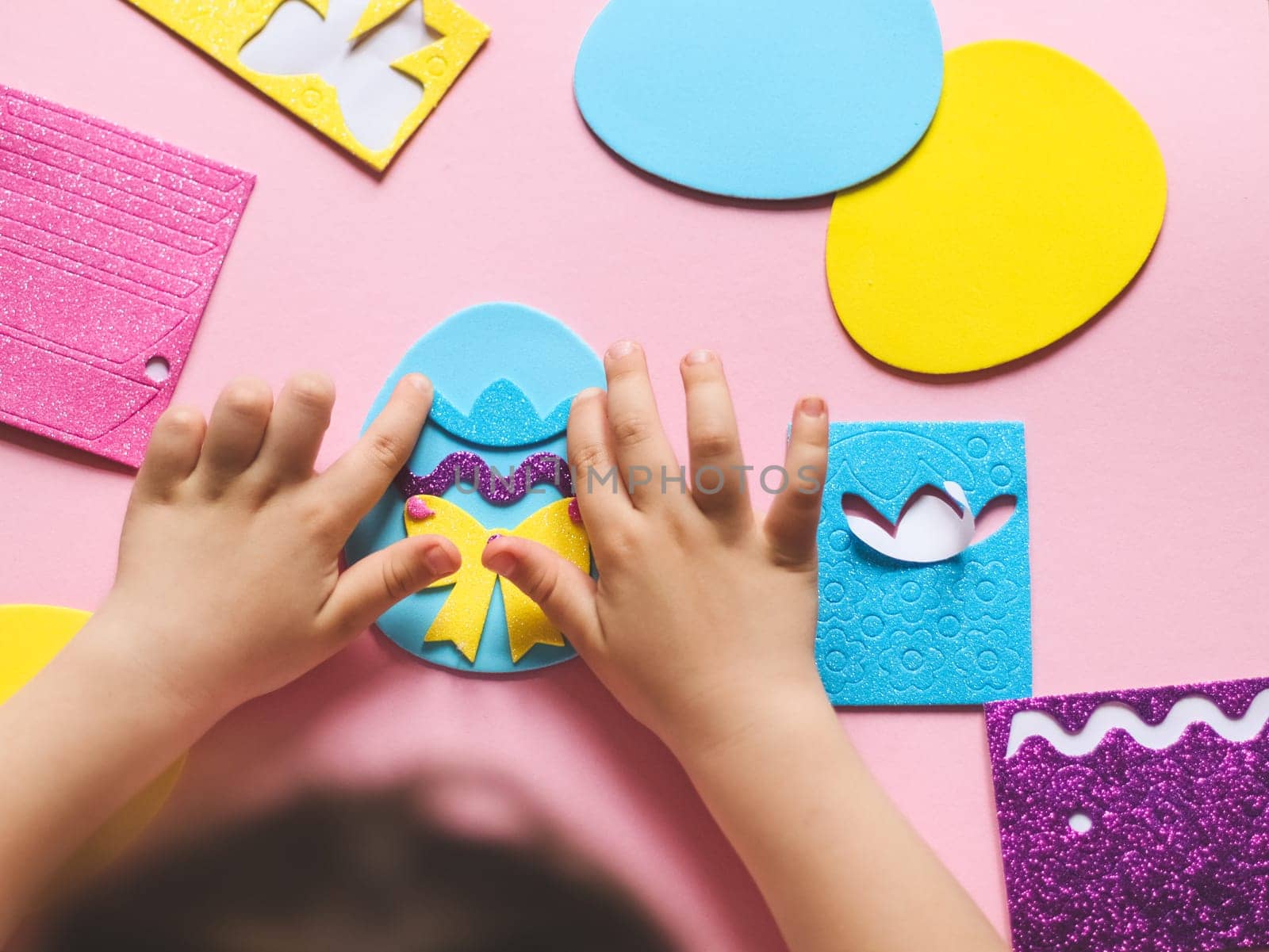 The hands of a little caucasian girl stick a hat sticker on a blue felt egg with a lilac zigzag by Nataliya
