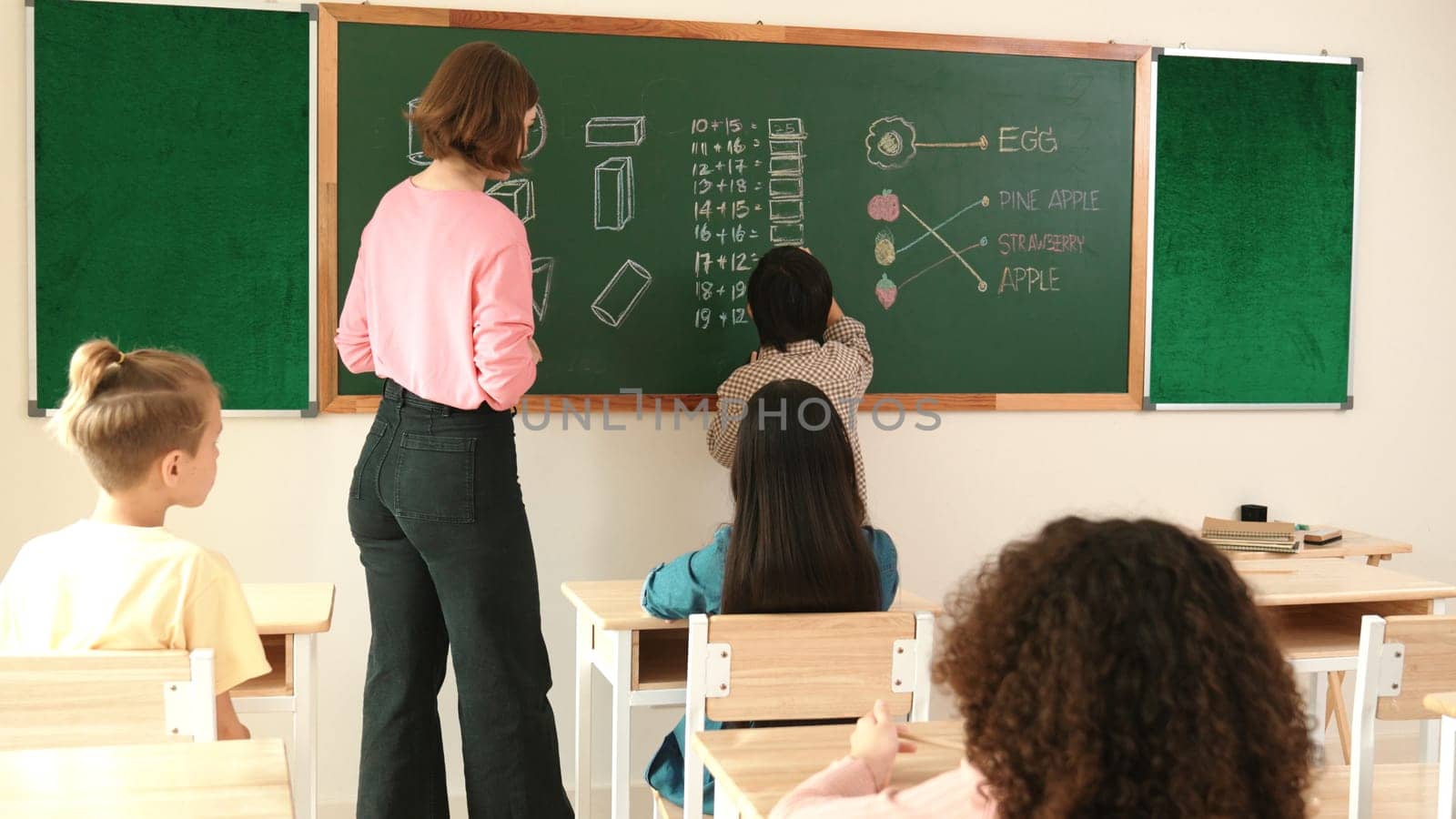 Smart teacher checking cute student homework while explaining idea at class. Attractive instructor walking around classroom while looking at notebook and talking about Math theory to child. Pedagogy.