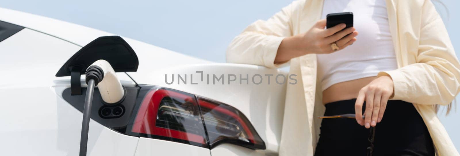 Young woman use smartphone to pay for electricity at public EV car charging station green city park. Modern environmental and sustainable urban lifestyle with EV vehicle. Panorama Expedient