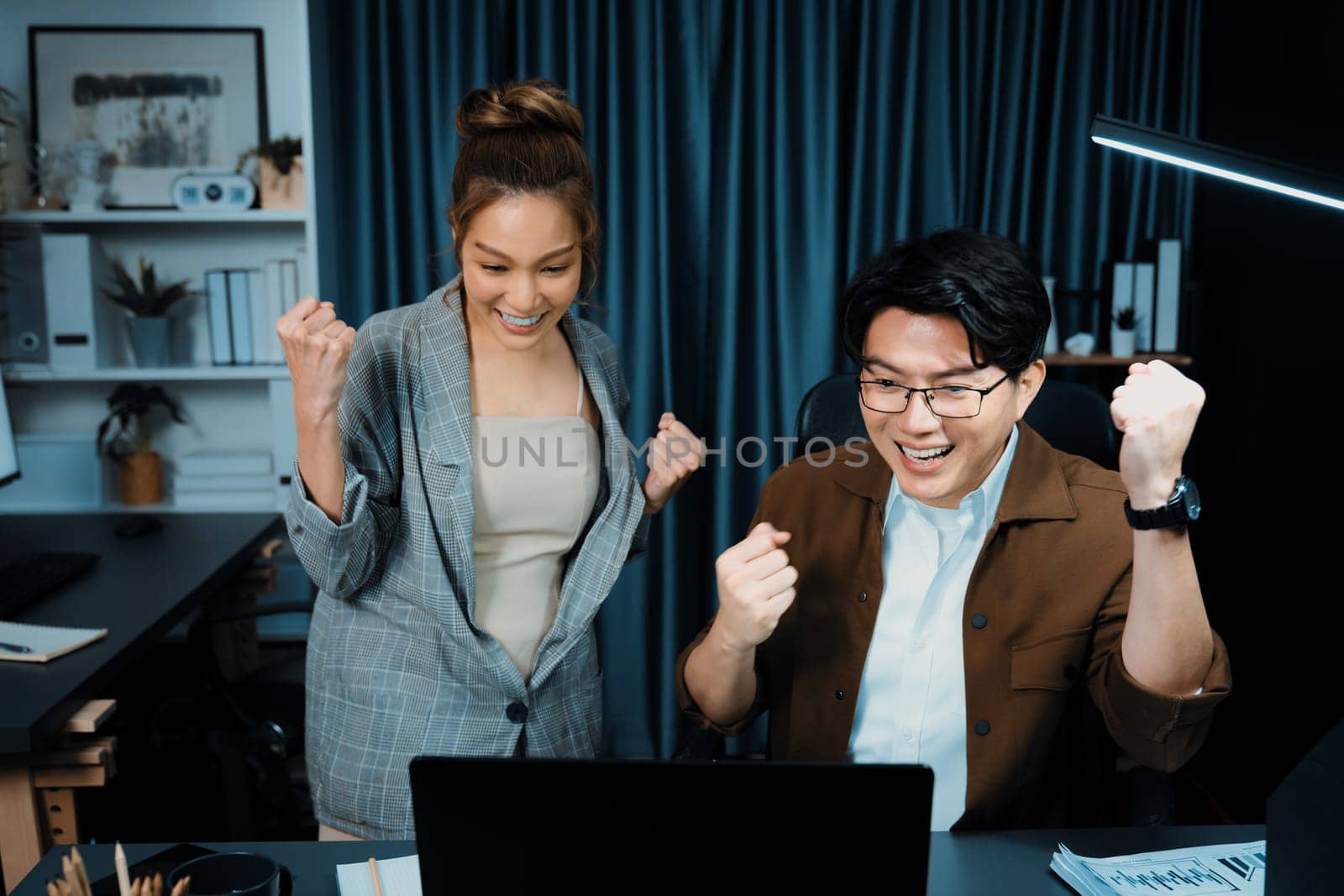 Successful smiling business partners earning high revenue in sales raising fist up in creative design architecture planning project job investment via email at modern home office at night. Infobahn.