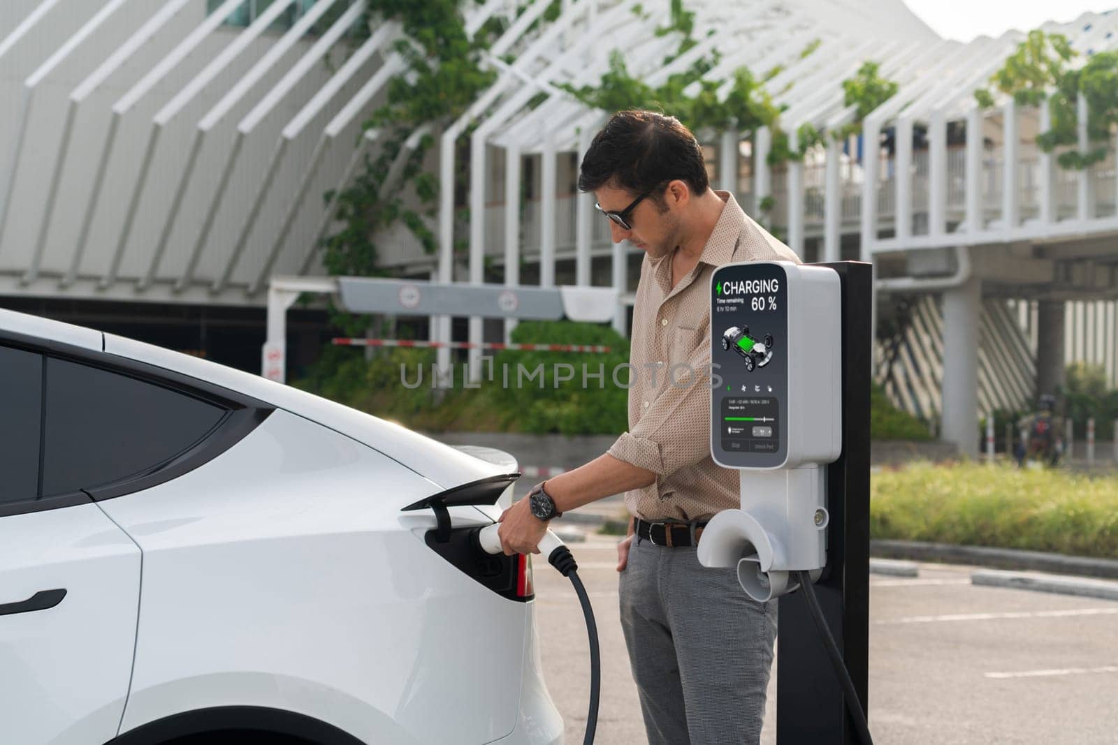 Young man put EV charger to recharge electric car's battery from charging station in city commercial parking lot. Rechargeable EV car for sustainable environmental friendly urban travel. Expedient