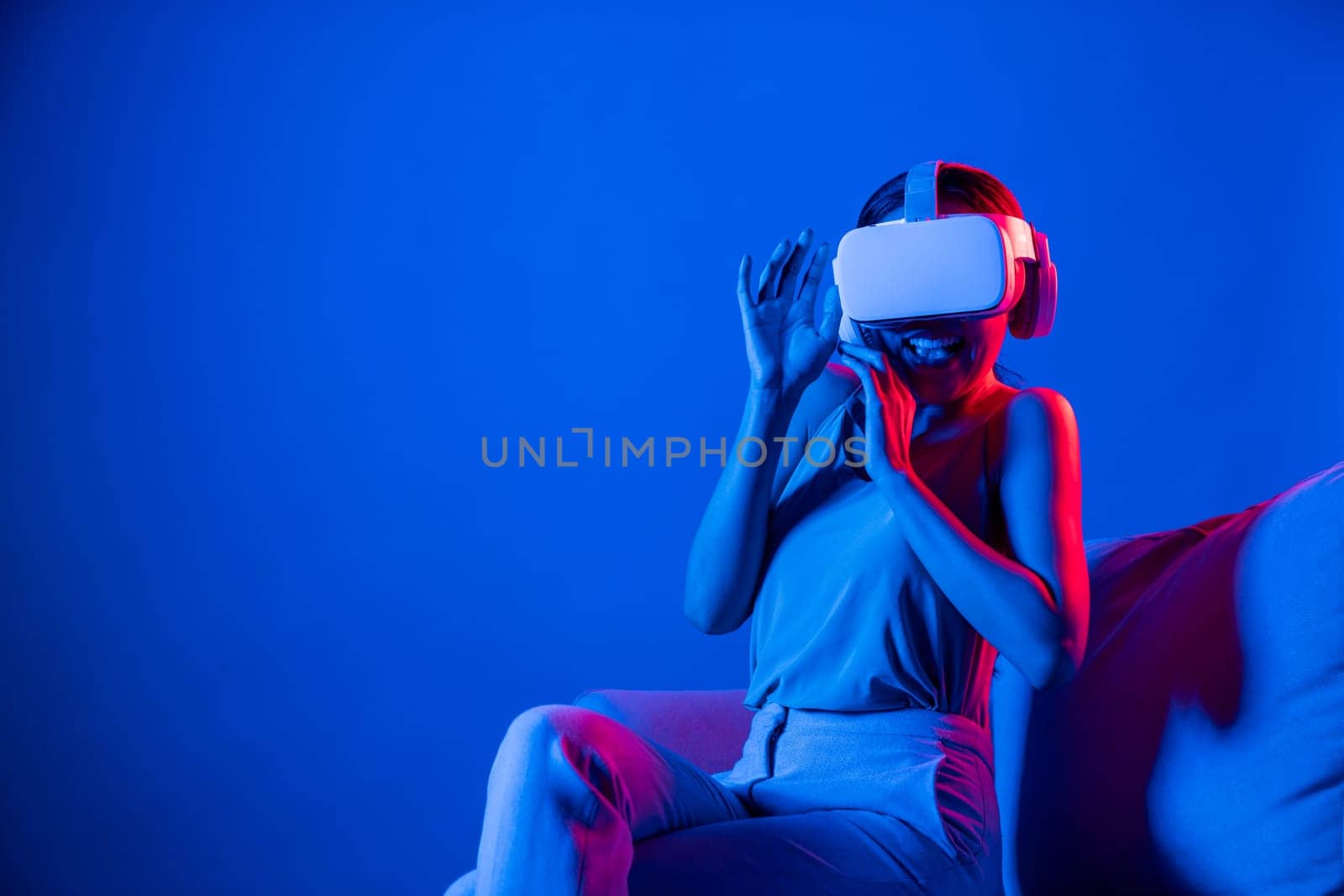 Smart female sitting on sofa surrounded by neon light wear VR headset connect metaverse, futuristic cyberspace community technology. Elegant woman excited and emotionally watch movie. Hallucination.