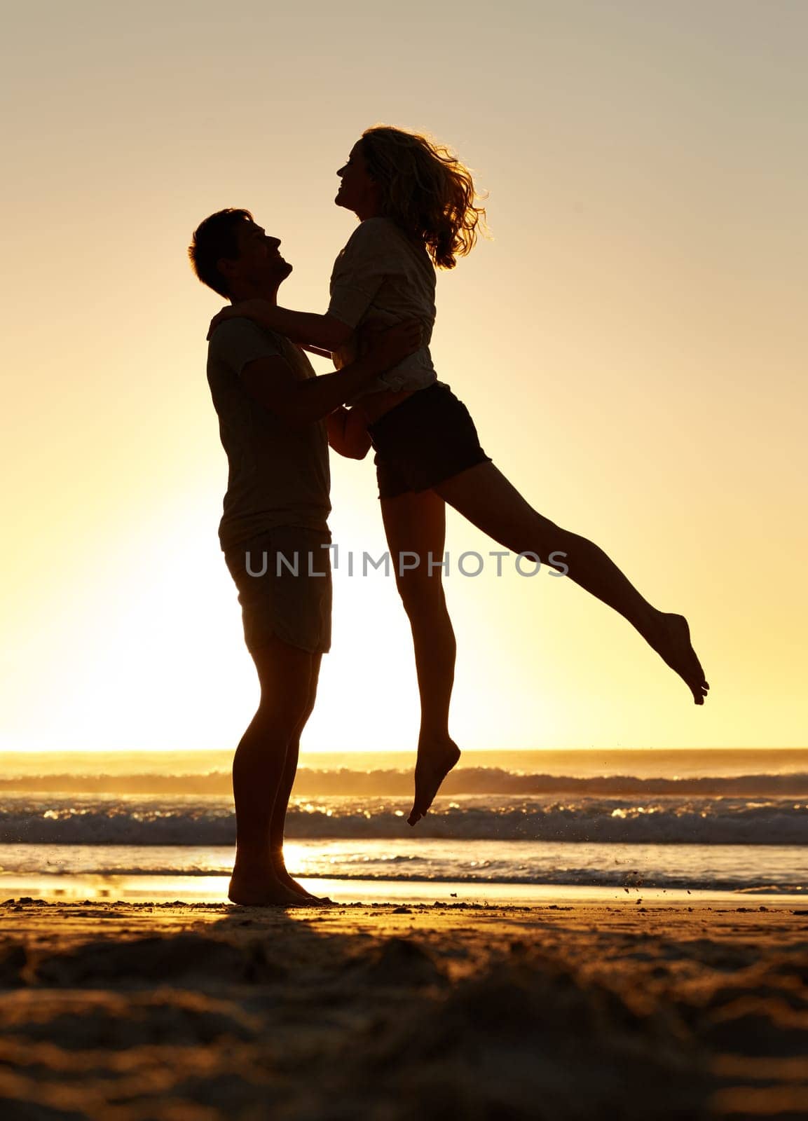 Lifting, silhouette and couple with sunset at beach, ocean and sea for affection, love and to relax. People, partners and sunrise for care together on vacation, holiday and travel in Australia.