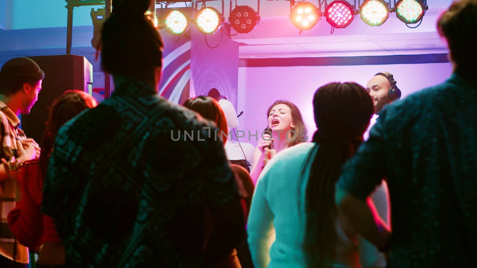 Happy woman doing karaoke at party, singing songs with DJ in front of friends on dance floor. Confident girl having fun with live show perfromance next to electronic mixing station at nightclub.