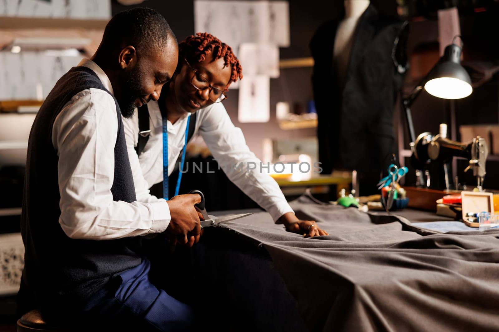 Experienced african american atelier shop coworkers meticulously cutting refined textile material for customer comissioned suit. Expert suitmakers manufacturing bespoke sartorial outfit